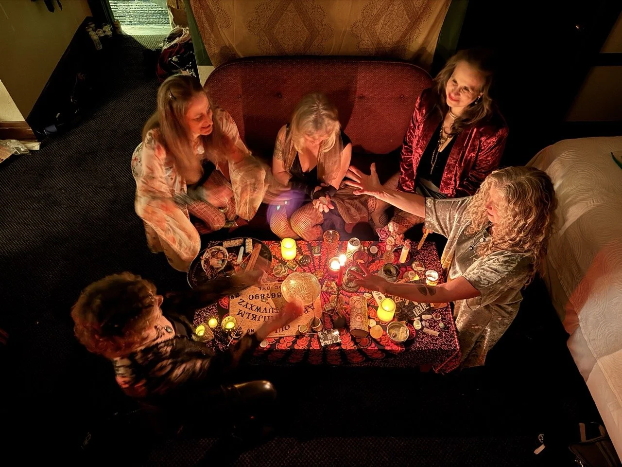 Five women gather around a table with candles, tarot cards, and crystals, in a dimly lit room, engaging in a tarot reading session.
