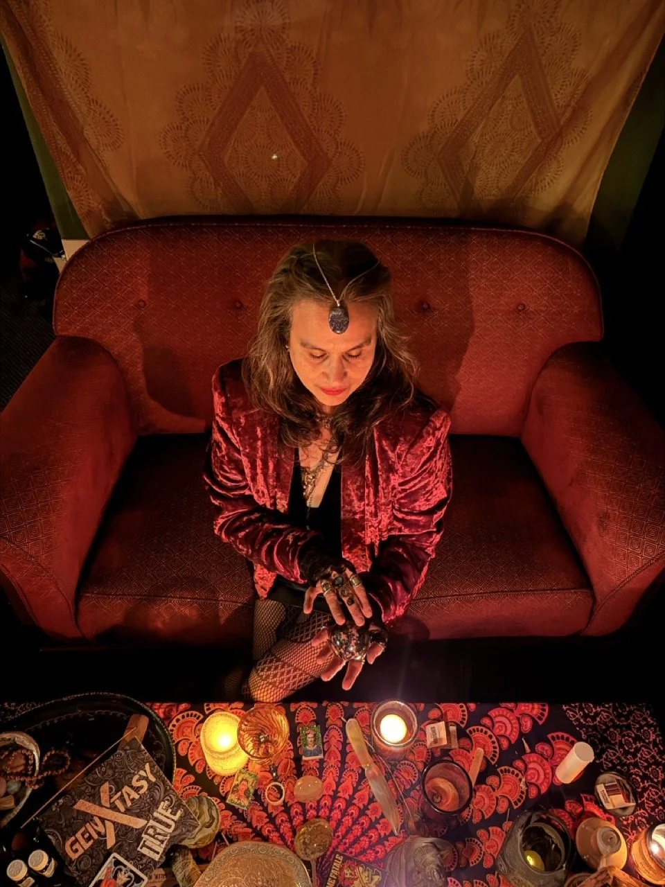 A woman sits on a red velvet sofa with her head bowed, wearing a red velvet jacket, fishnet stockings, and gothic jewelry. There are candles and various objects on a table in front of her, creating a mystical or ritualistic atmosphere.
