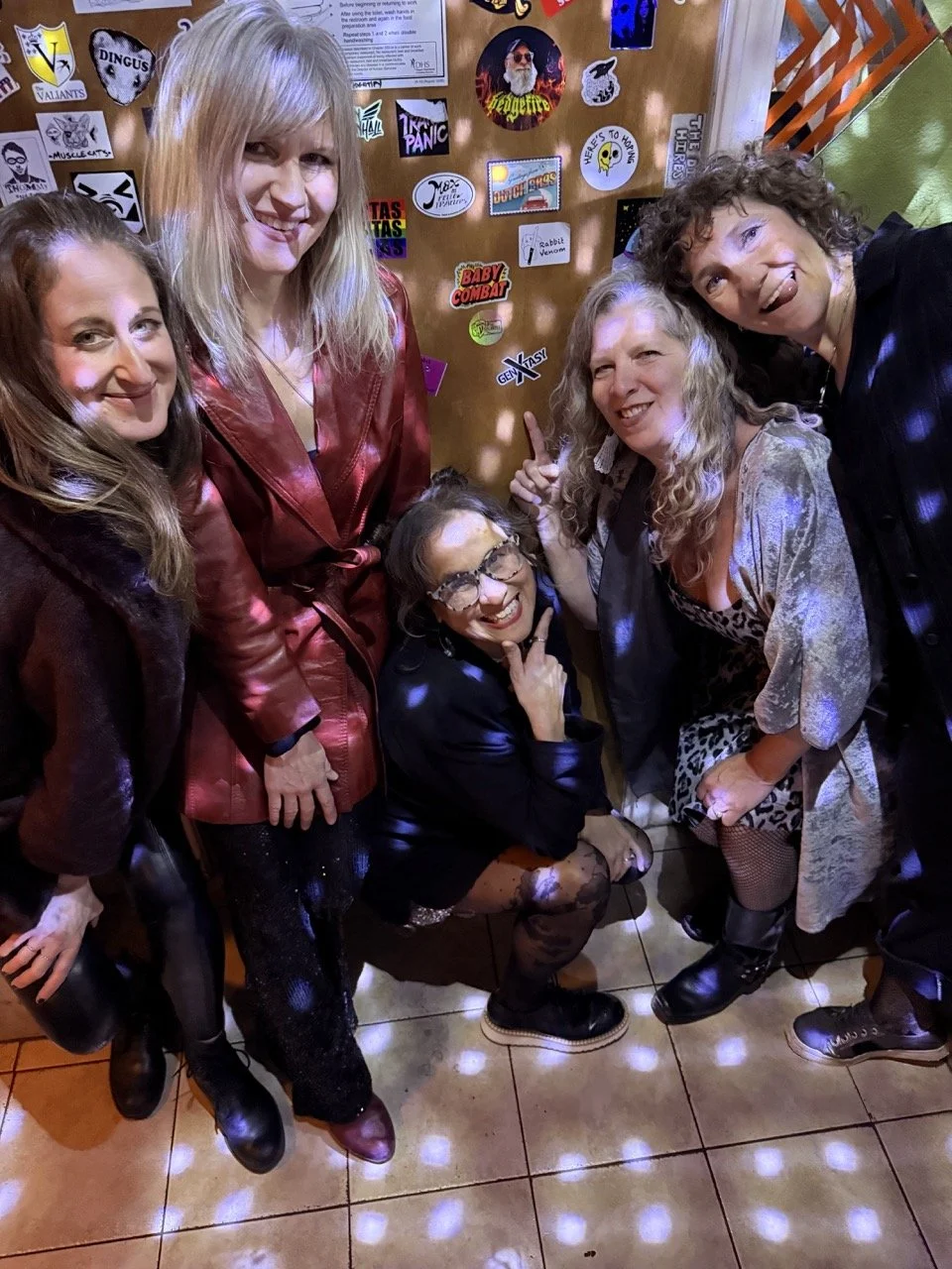 Group of five women posing together indoors, some smiling, some making playful gestures. The background features a decorated wall with various stickers and signs. The women are dressed in casual and party attire.