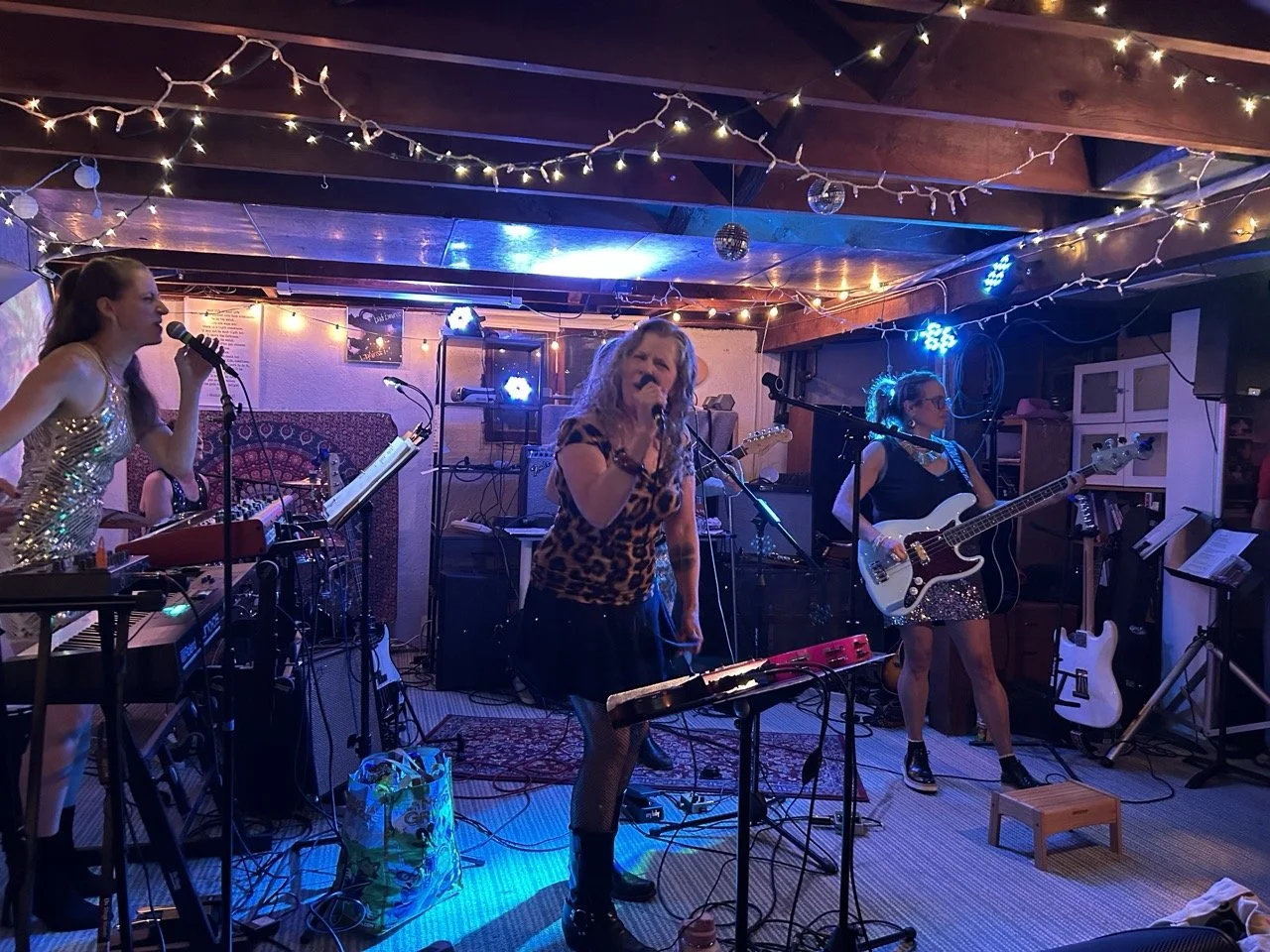 A band of five female musicians performing in a cozy, decorated room with string lights and a disco ball.