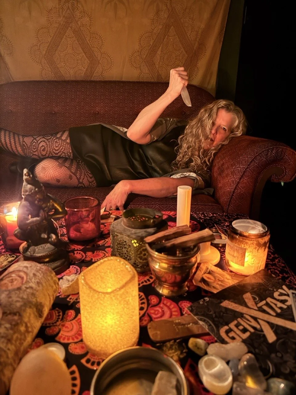 A woman with curly blonde hair lying on a red patterned sofa, holding a large knife, surrounded by lit candles, figurines, and various objects on a table in a cozy, dimly lit room.