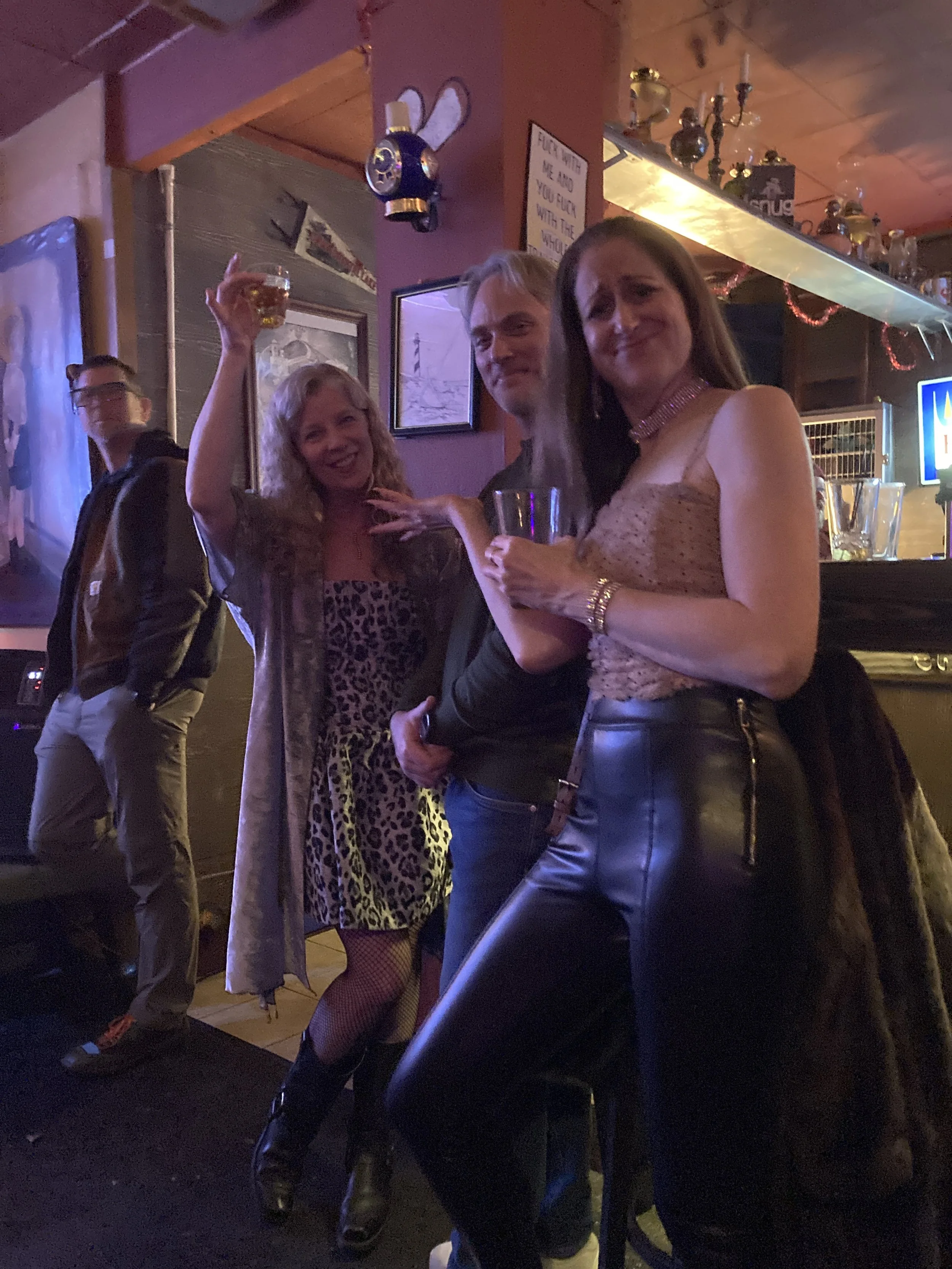 Group of people socializing in a bar, holding drinks, smiling, with a bartender in the background, dim lighting, and bar decor.
