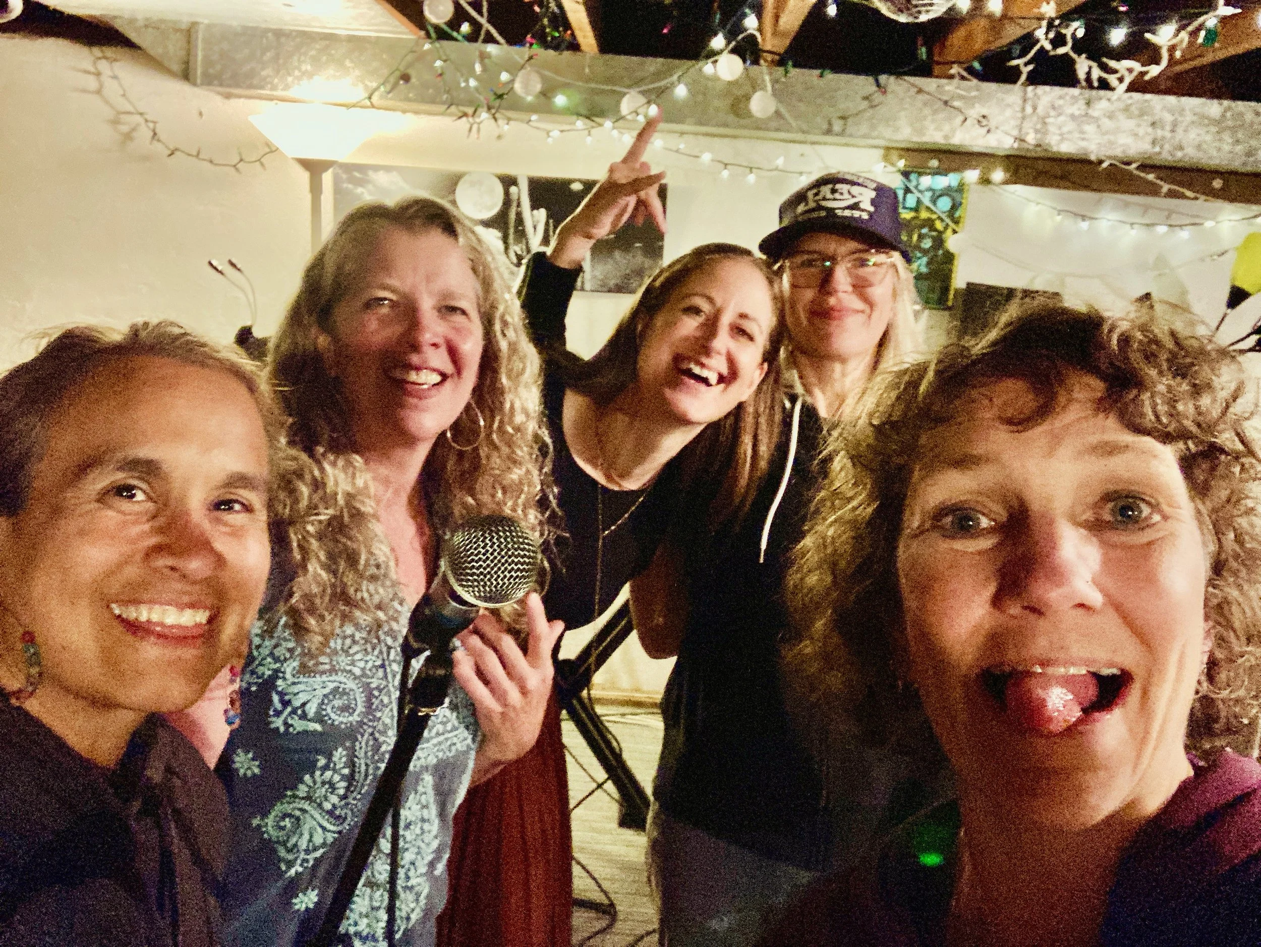 Group of six women taking a selfie at a party with string lights and decorations in the background. One woman is holding a microphone, and the woman at the front is sticking out her tongue.