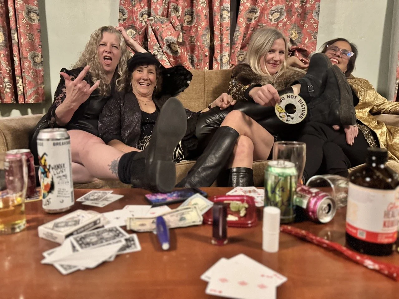 Four women sitting on a couch with drinks and scattered playing cards on the table in front of them, casually posing and smiling.