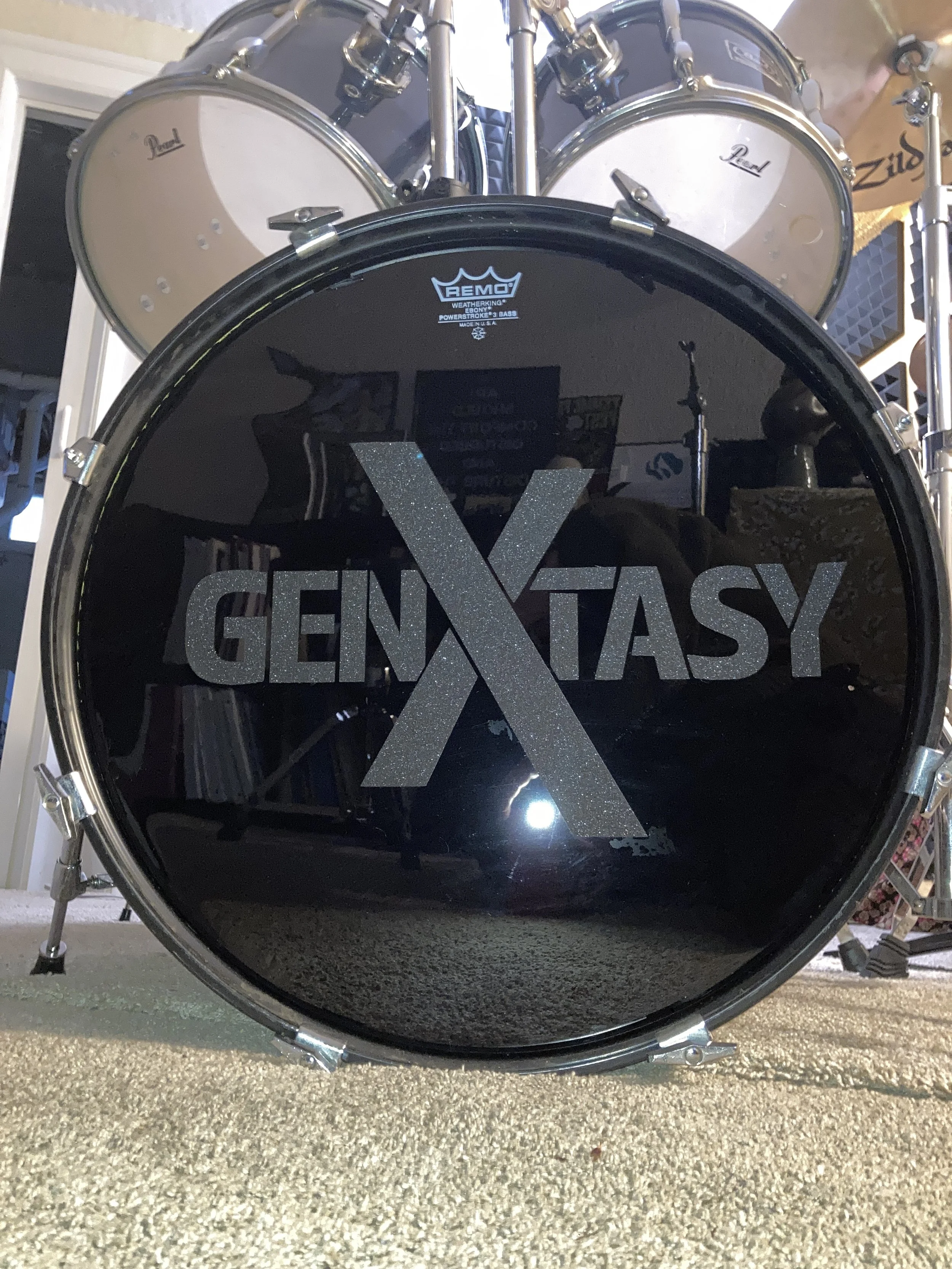 Close-up of a black drum with glittery text reading ‘GENT X TASY’ on the front, with a reflection of a person and a room in the drum's surface, and a four-piece drum set in the background.