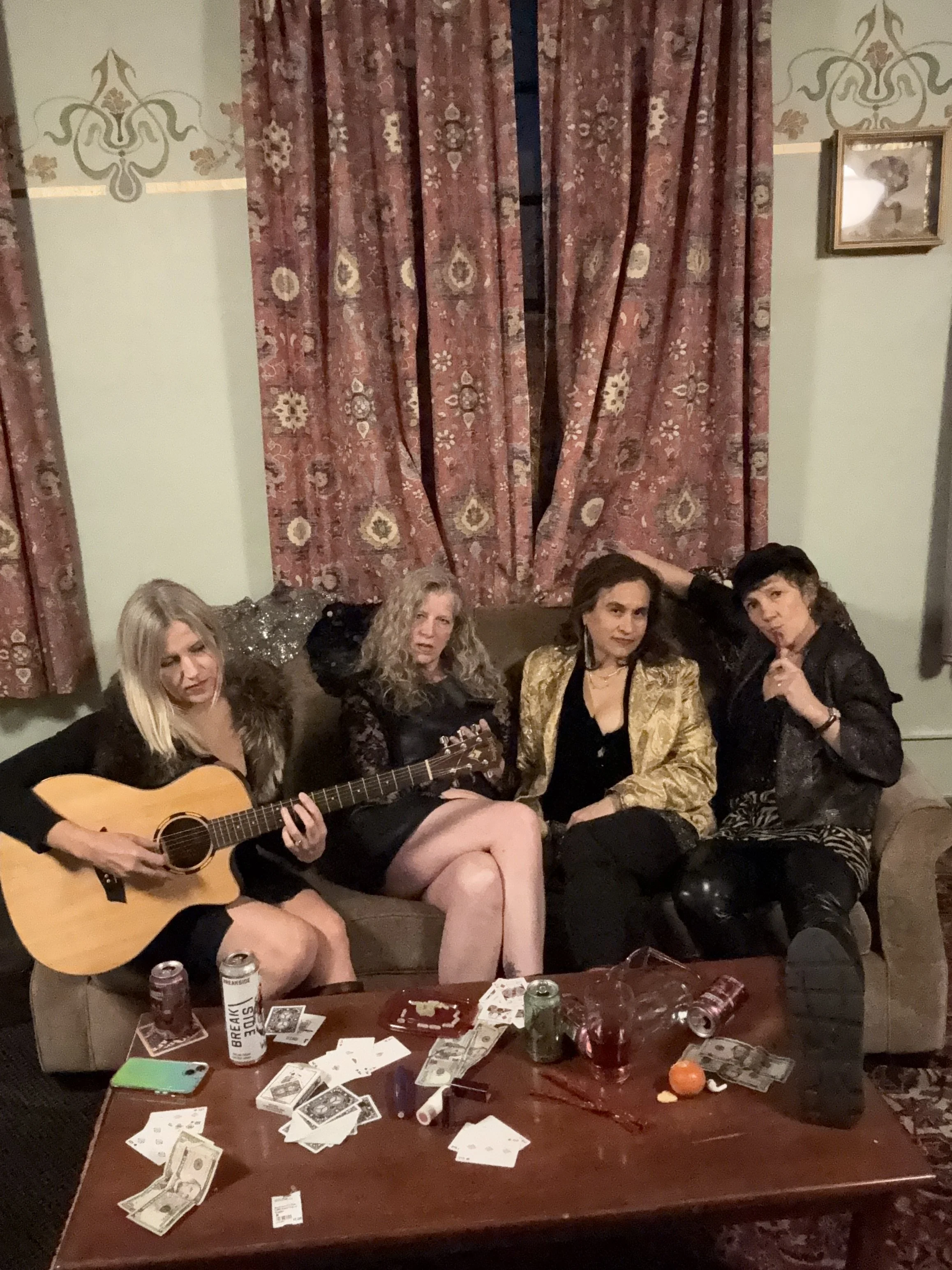 A group of four women sitting on a sofa in a living room with pink patterned curtains. One woman is playing a guitar, and the others are posing. The coffee table in front has money, drinks, and snacks, creating a party atmosphere.