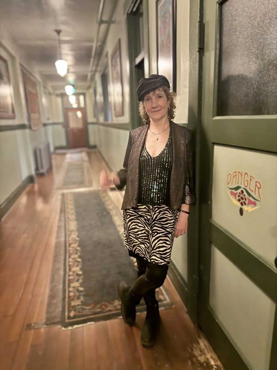A woman standing in a hallway wearing a black beret, sparkly top, animal print skirt, black tights, and boots, leaning against a door with a 'DANGER' sign on it.