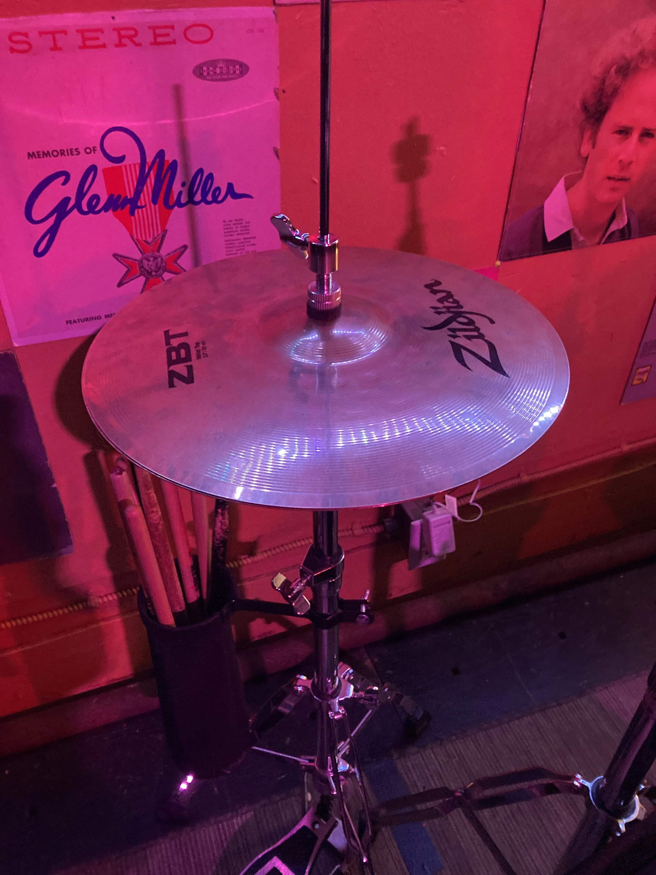 A drum cymbal mounted on a stand, with pink lighting, positioned in front of record album covers featuring Glenn Miller and Billy Joel in the background.
