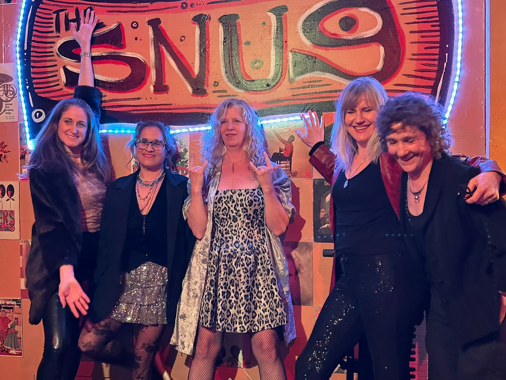 Six women dressed in vintage and glamorous clothing, posing and smiling in front of a brightly colored sign reading 'The Swing' at a lively event or party.