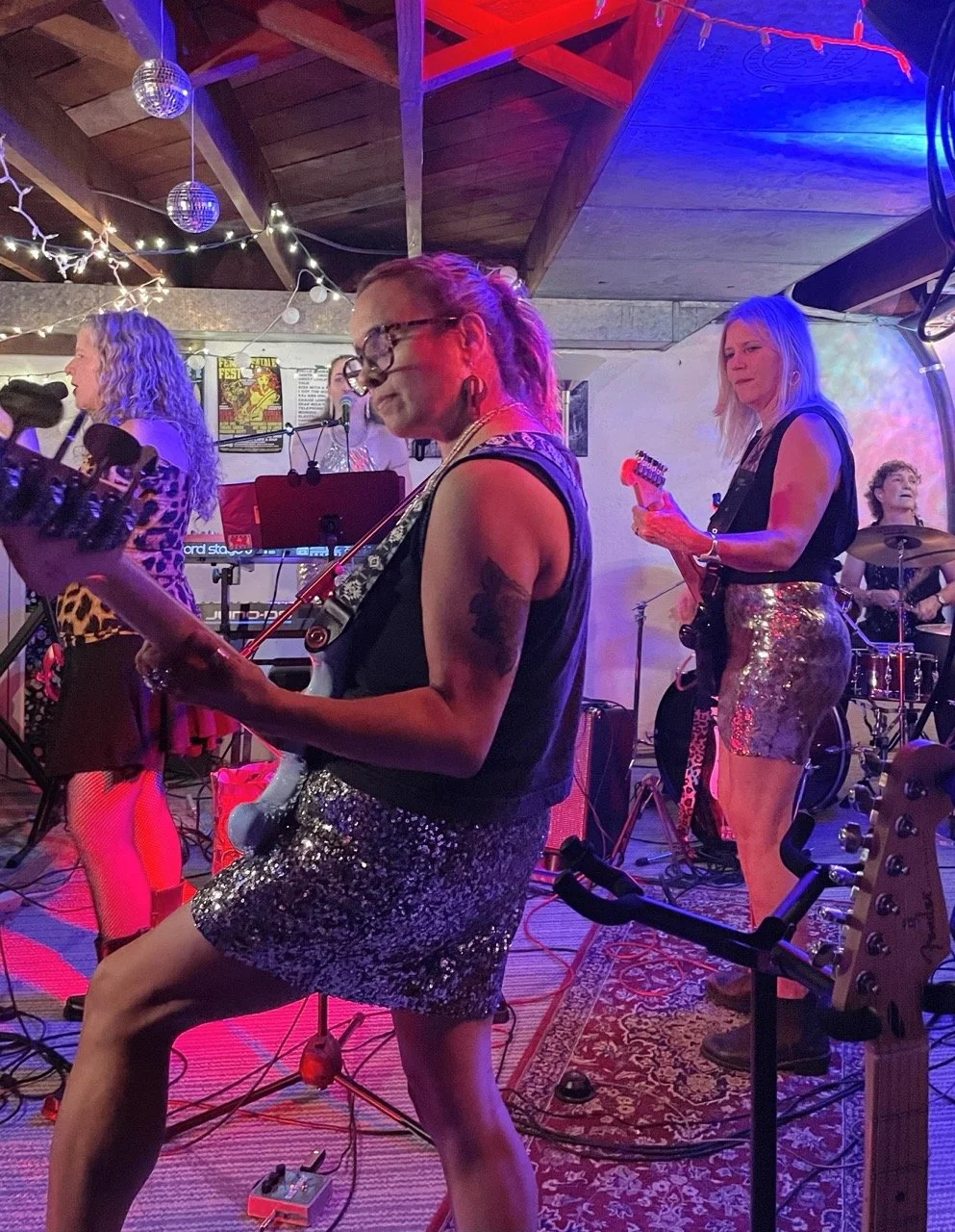 A band of women performing on stage, with colorful lighting, musical instruments, and a decorated indoor venue.