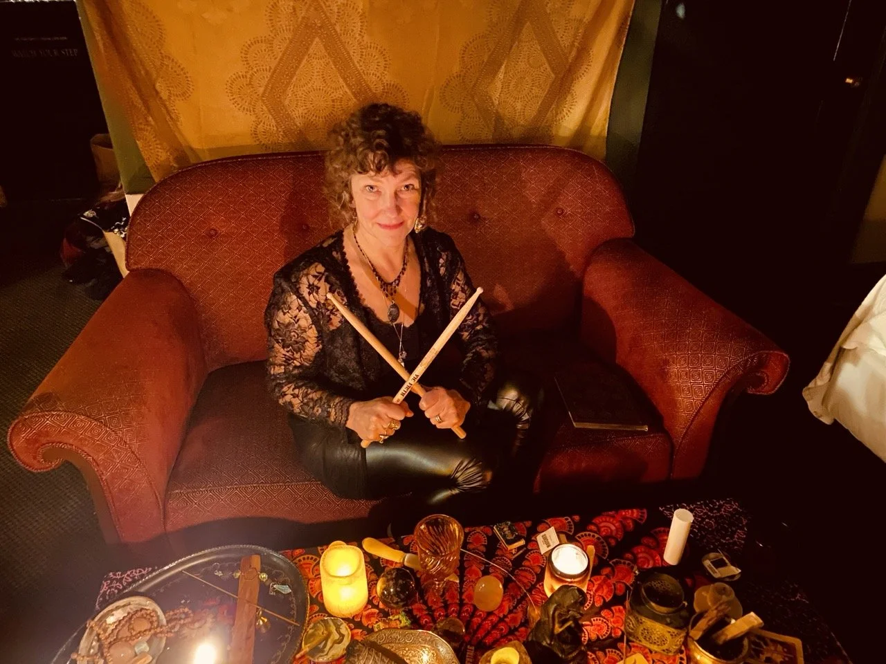 A woman with curly hair wearing a black lace top and leather pants, sitting on a red vintage sofa, holding two drumsticks crossed in her hands. There are candles, crystals, and various decorative items on the table in front of her, suggesting a mysti