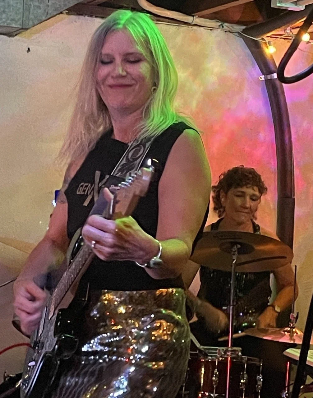 Two women performing music with a woman in front playing an electric guitar and another woman behind on drums, colorful lights behind them.