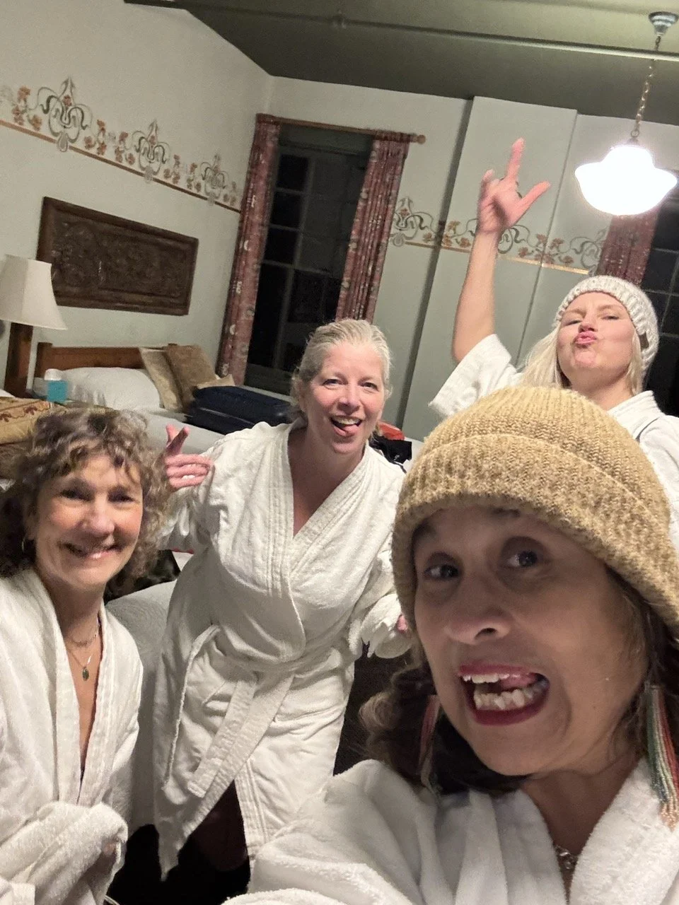 Four women in bathrobes posing for a selfie in a cozy, vintage-style bedroom with floral wallpaper, red curtains, and warm lighting.