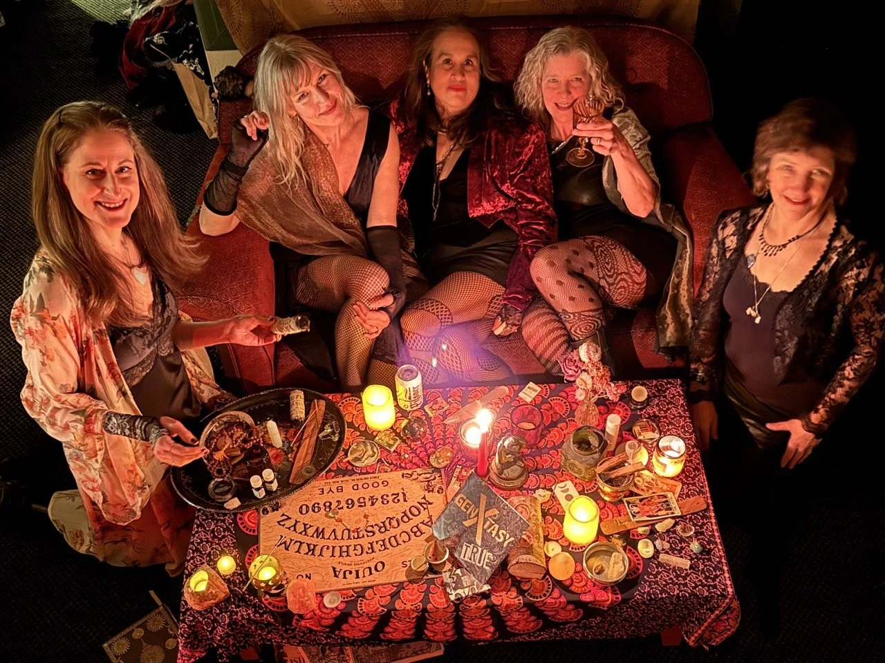 Five women sitting and standing around a table decorated for a séance, with candles, tarot cards, and mystical items, at a party or gathering.
