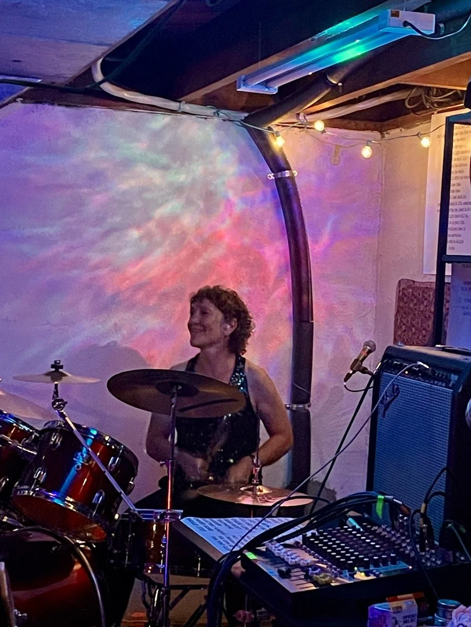 A woman with short curly hair playing drums in a dimly lit room with colorful lighting and a nebula-patterned backdrop.