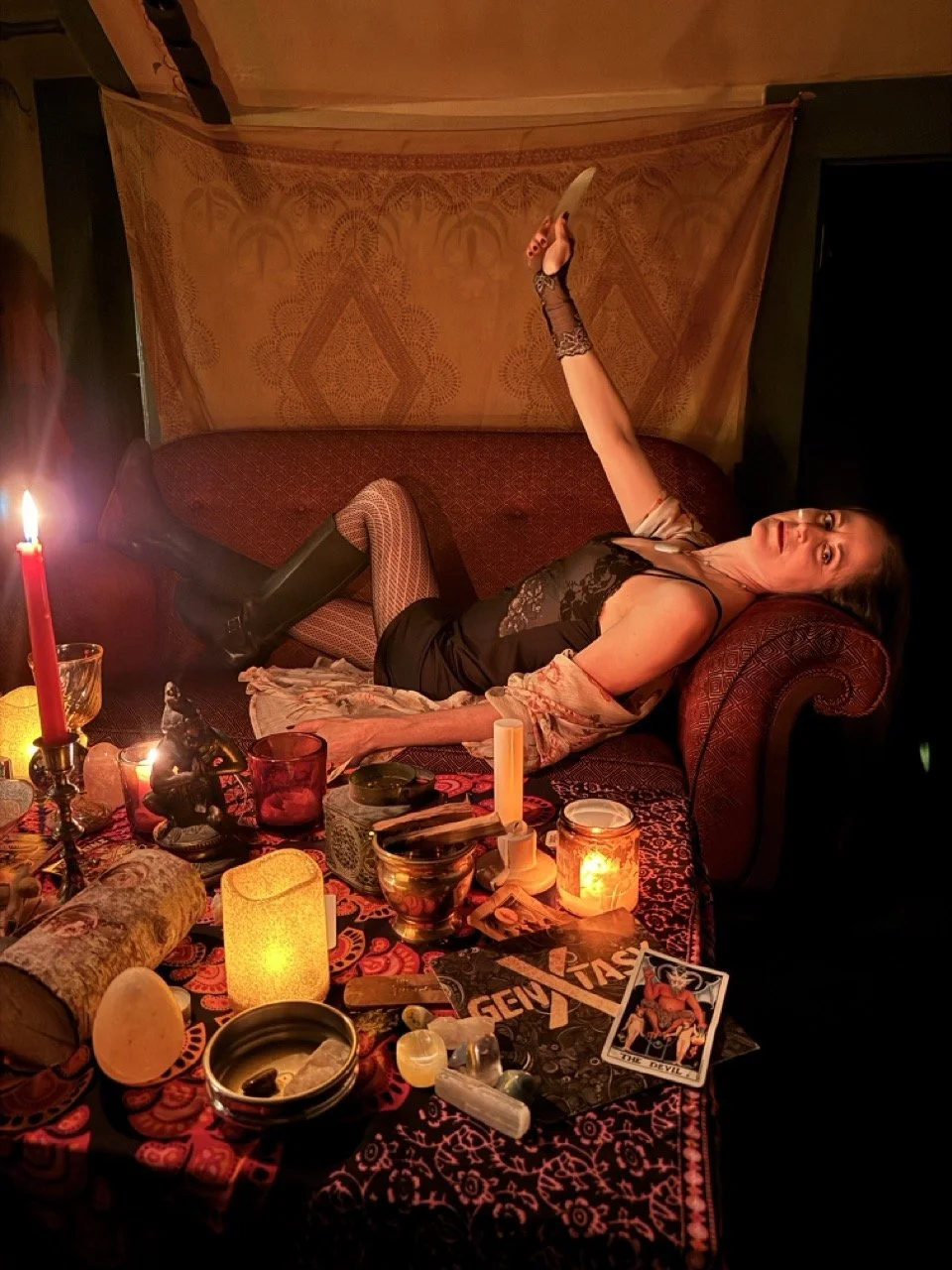 A woman lying on a sofa with one leg raised, surrounded by candles, tarot cards, and mystical objects in a dimly lit room.