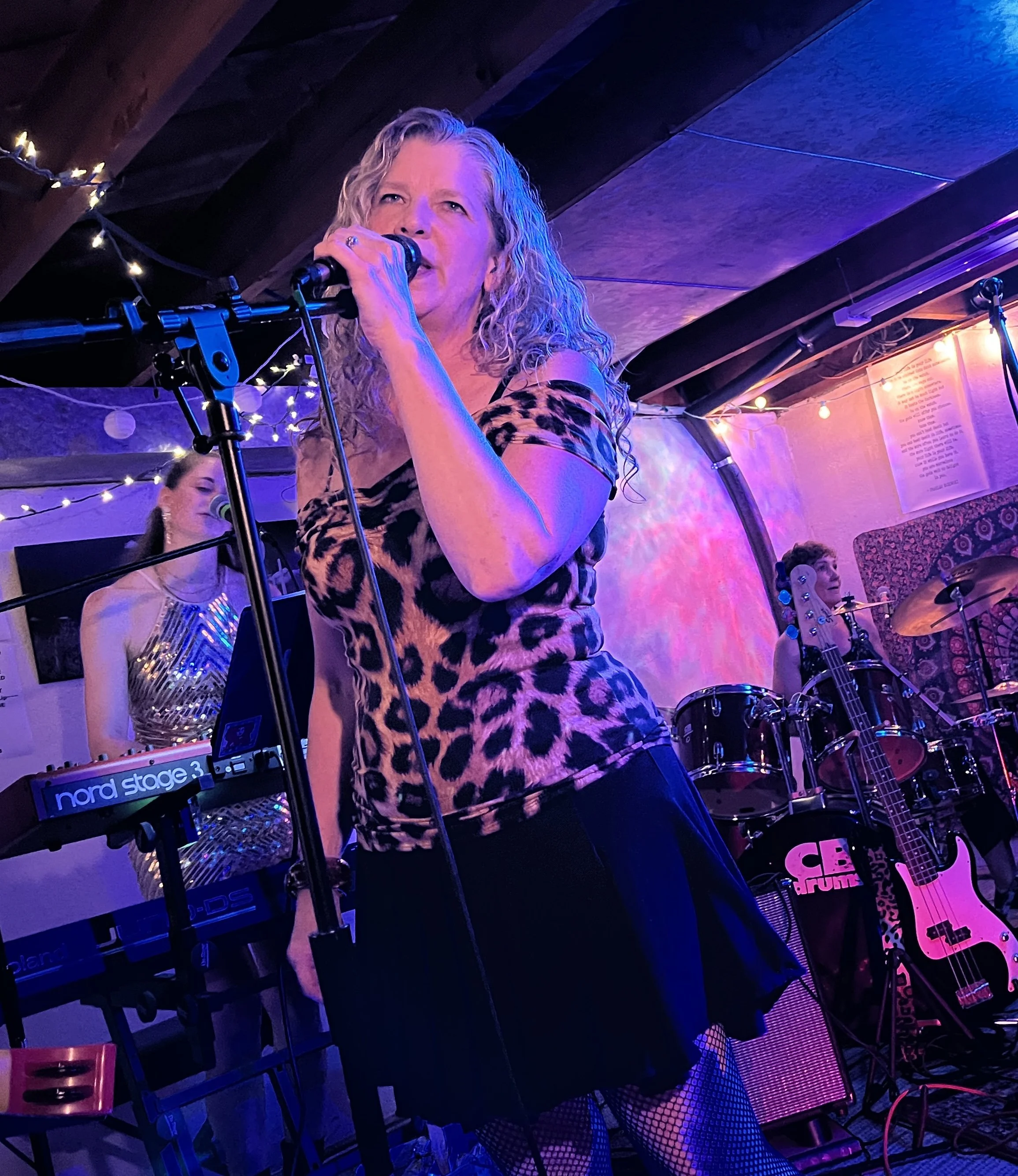 A woman sings into a microphone on stage with a leopard print top, with another woman playing keyboard and a third woman on drums in the background, all under colorful stage lighting.