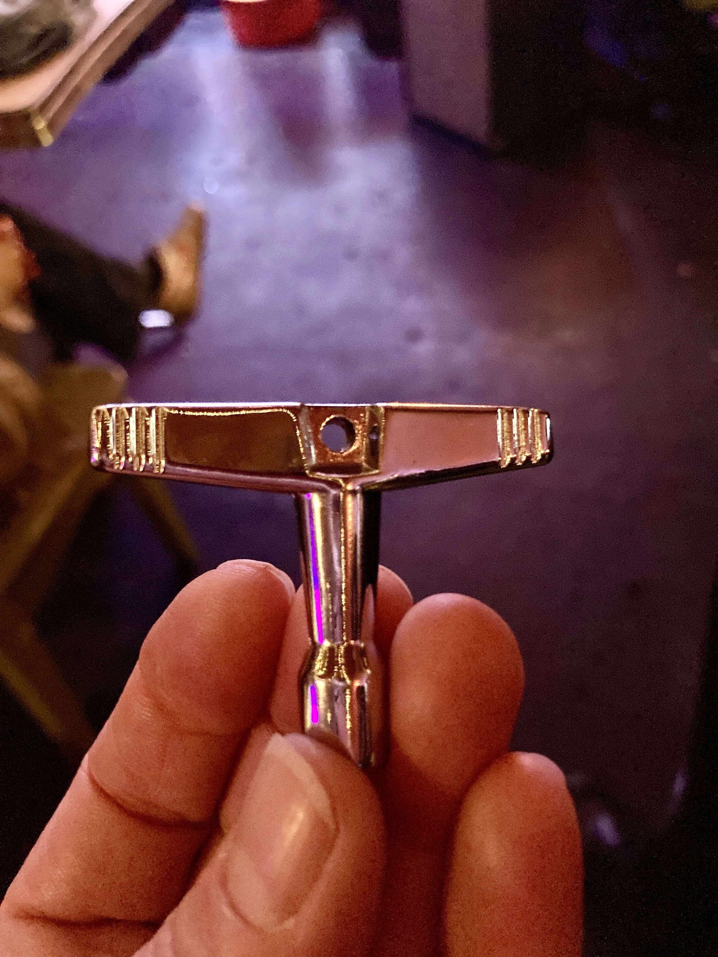 Close-up of a hand holding a vintage safety razor with a brass-colored handle and a flat, wide head, against a dimly lit room with a wooden floor and furniture.