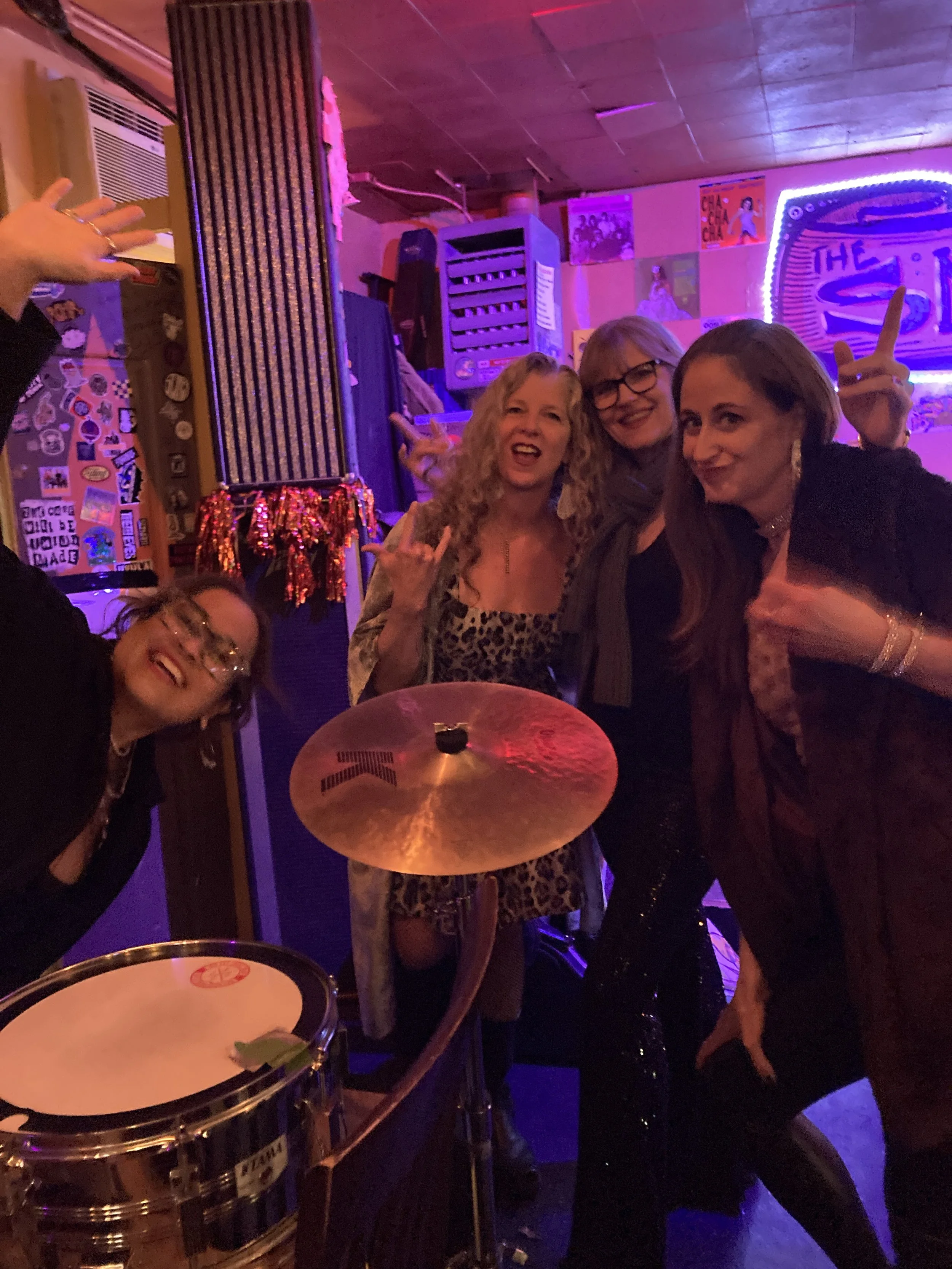 Four women having fun at a lively, colorful bar or club with neon purple and pink lighting, musical instruments, and various posters and decorations on the walls.