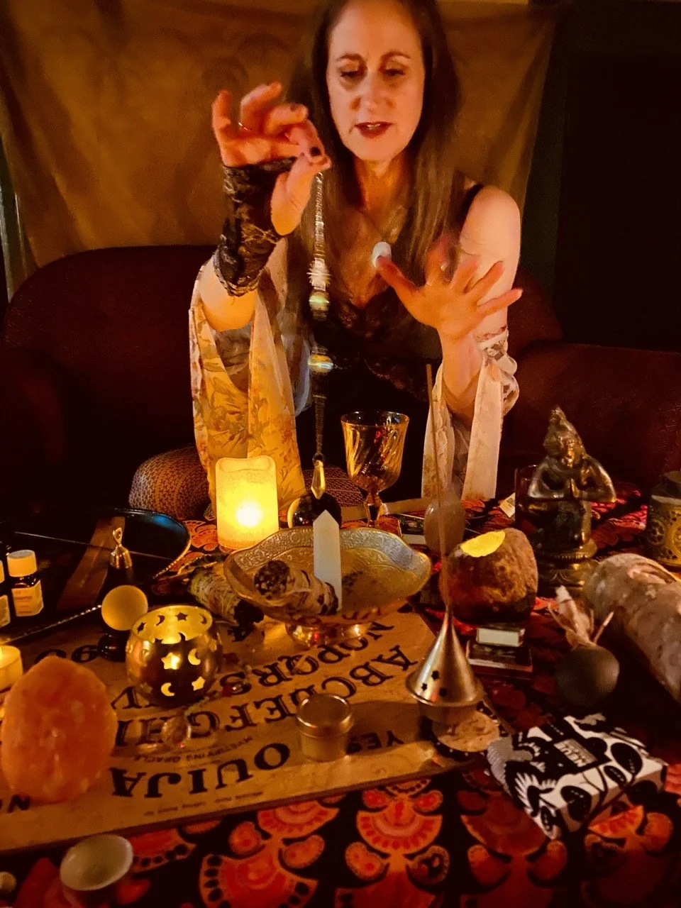 A woman with long brown hair wearing a black top and lace shawl, standing behind a table with candles, crystals, and mystical items, holding an amulet or charm in her hand, in a dimly lit room.