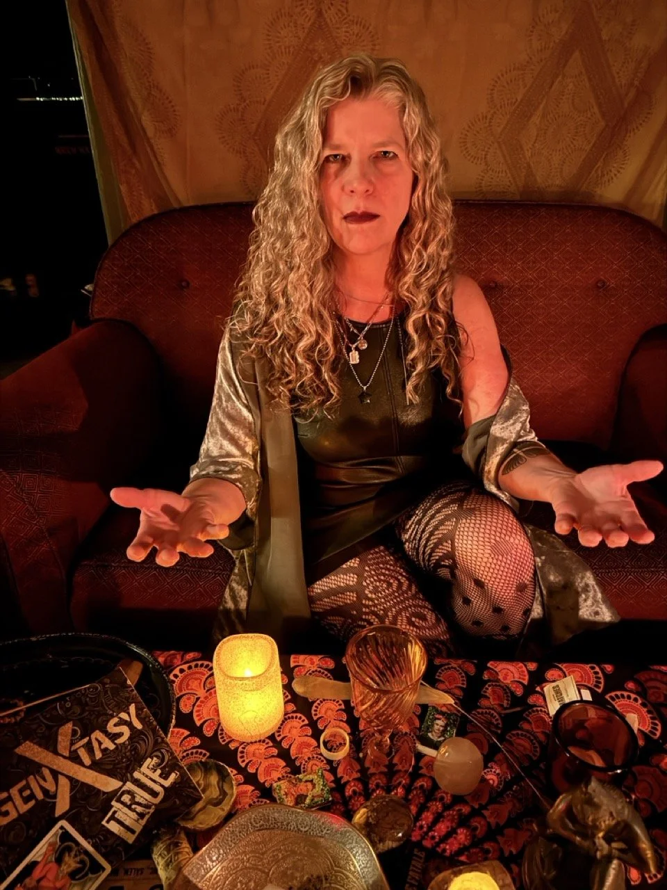 A woman with long curly blonde hair, sitting on a patterned couch in a dimly lit room with a red glow, surrounded by candles, tarot cards, and decorative objects, making a questioning gesture with her hands.
