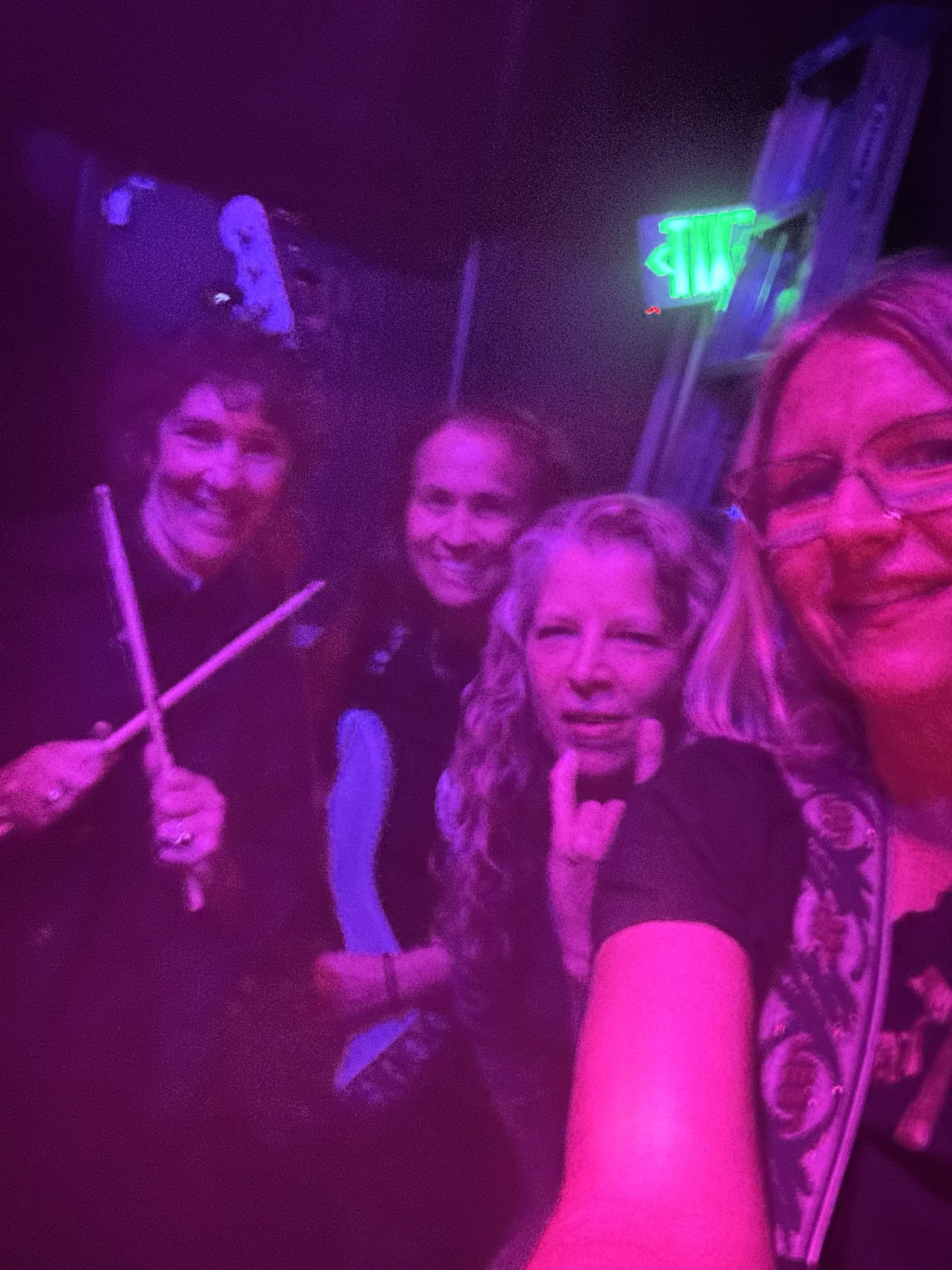 Four women taking a group selfie in a dimly lit room with purple and pink lighting, a neon green exit sign, and some posters on the wall.