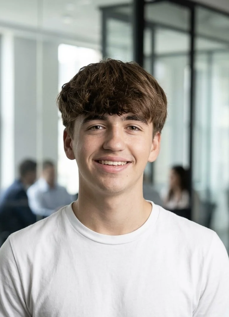A young man with brown, curly hair smiling, wearing a white t-shirt, in a modern office setting with glass walls and other people in the background.
