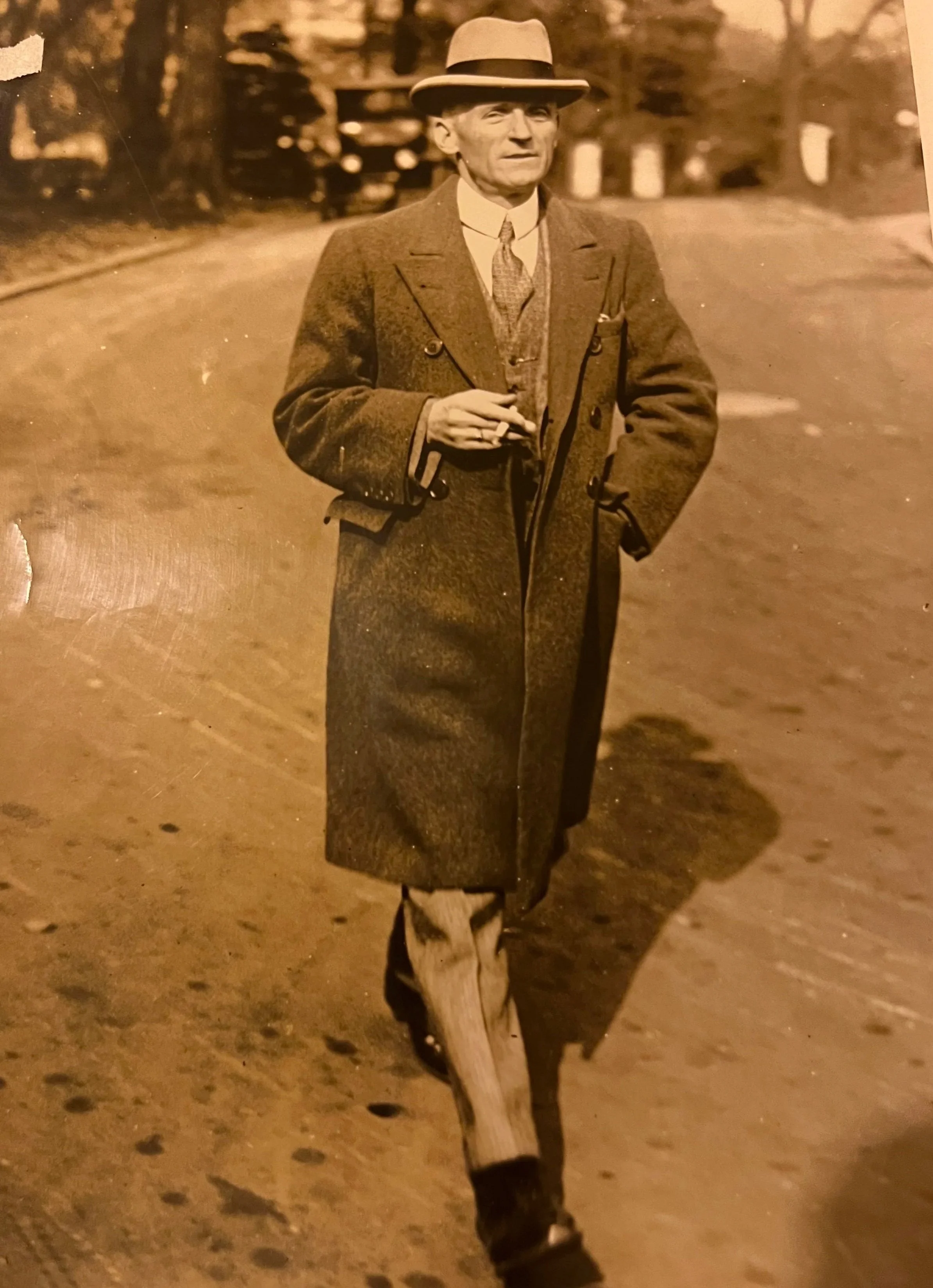 A sepia-toned photograph of a man walking outdoors on a street. He is dressed in vintage clothing, including a long coat, vest, tie, and a fedora hat. He is holding a cigarette in one hand and has one hand in his coat pocket.