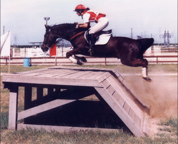 A person in a red and white helmet and riding gear riding a horse over a wooden jump at a racetrack.