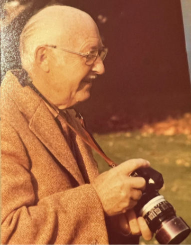 An elderly man with glasses and white hair, wearing a brown jacket, is smiling and holding a camera outdoors.
