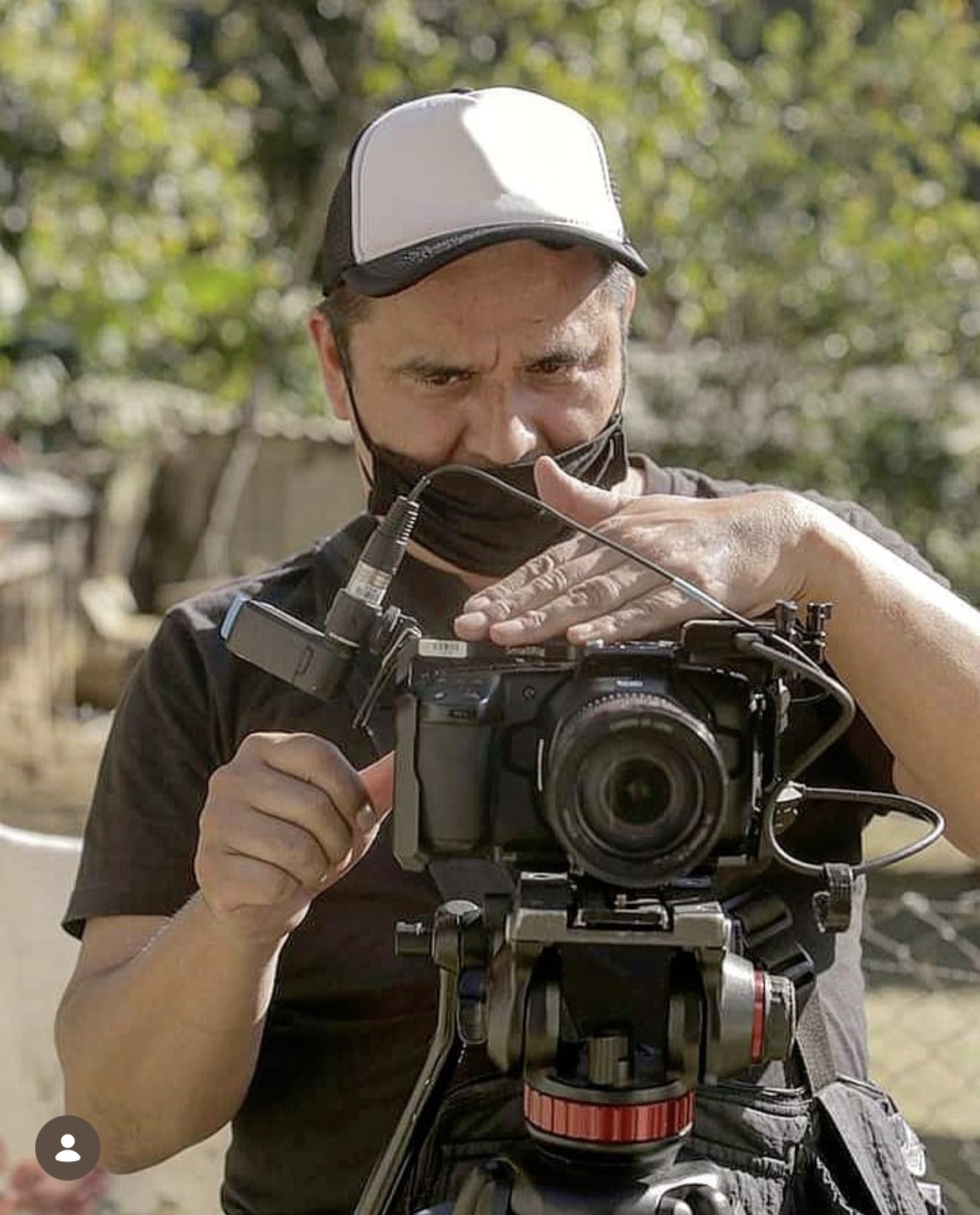 A man wearing a white and gray cap and a black mask is adjusting his camera mounted on a tripod outdoors with green trees in the background.