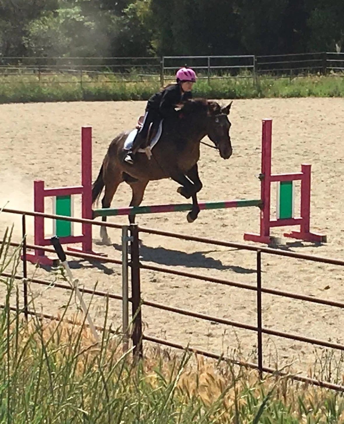 A young girl riding a brown horse over a jump in an outdoor equestrian arena, wearing a pink helmet and black riding outfit.
