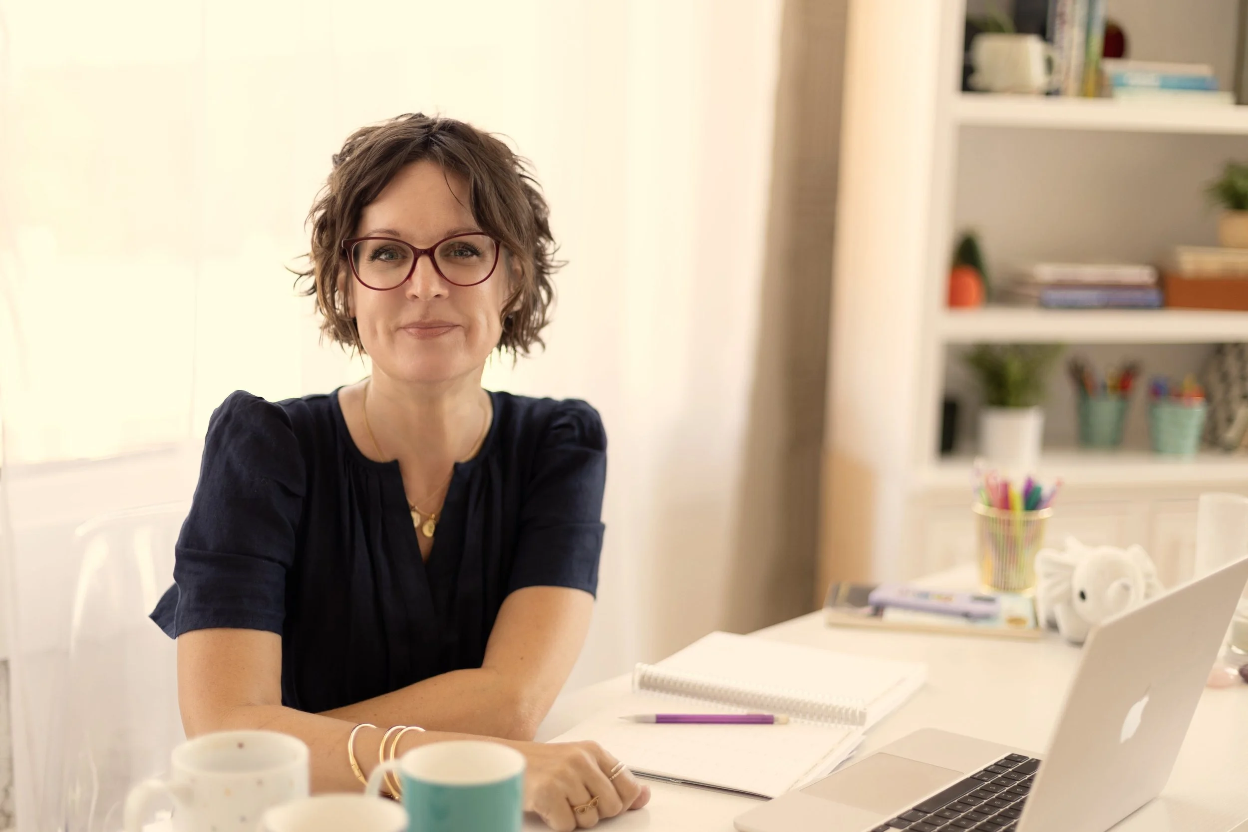 Ann Abbey, with short, wavy brown hair and glasses sits at a white table with notebooks, pens, and mugs, in a well-lit room with shelves of books and plants in the background.