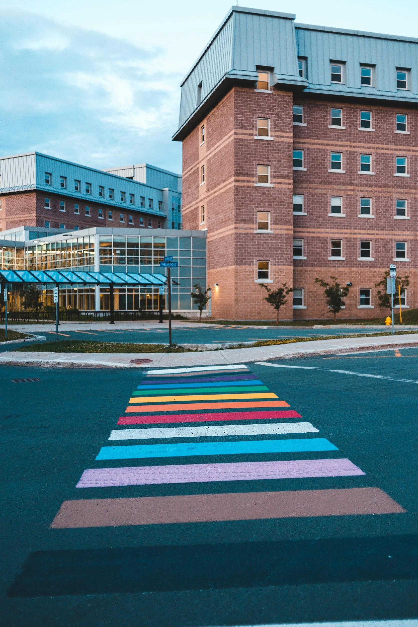 Colorful LGBTQIA+ rainbow crosswalk in front of a modern brick and glass building under a partly cloudy sky.