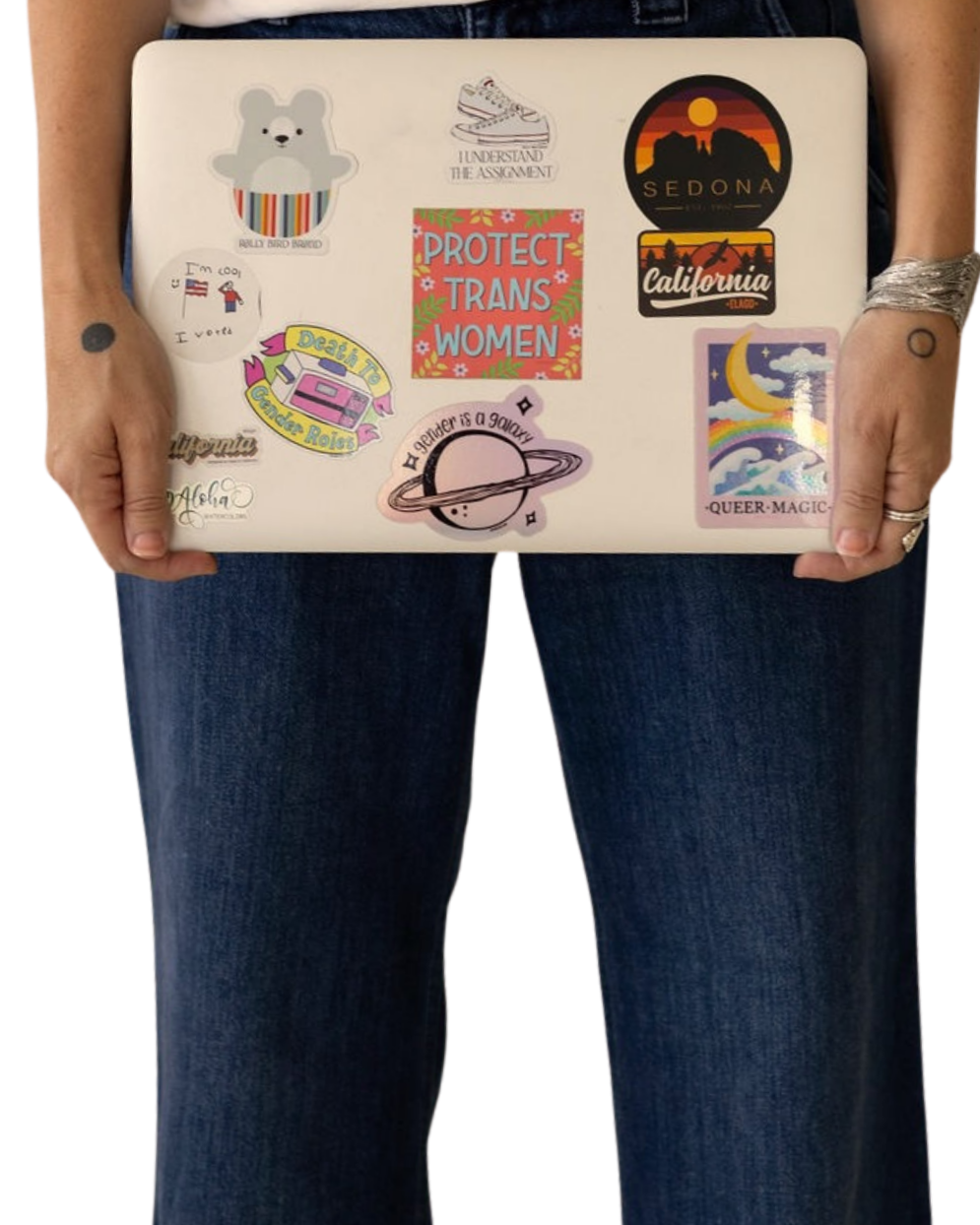 Person holding a laptop decorated with various stickers including a bear, rainbow, planets, and slogans supporting LGBTQ+ and social causes, wearing blue jeans and a bracelet.