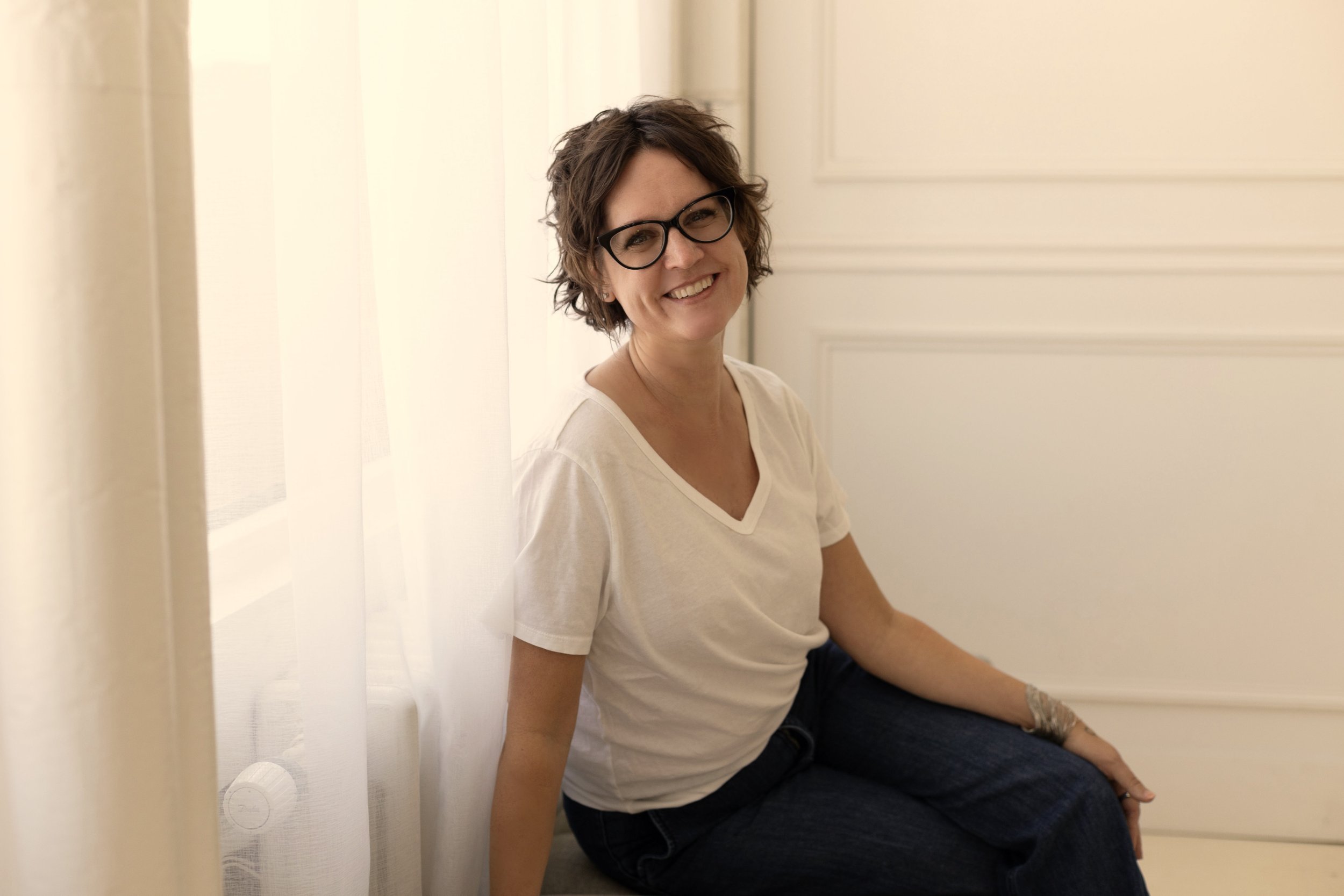Woman with short curly hair and glasses sitting on the floor and smiling inside a room with white walls and curtains.