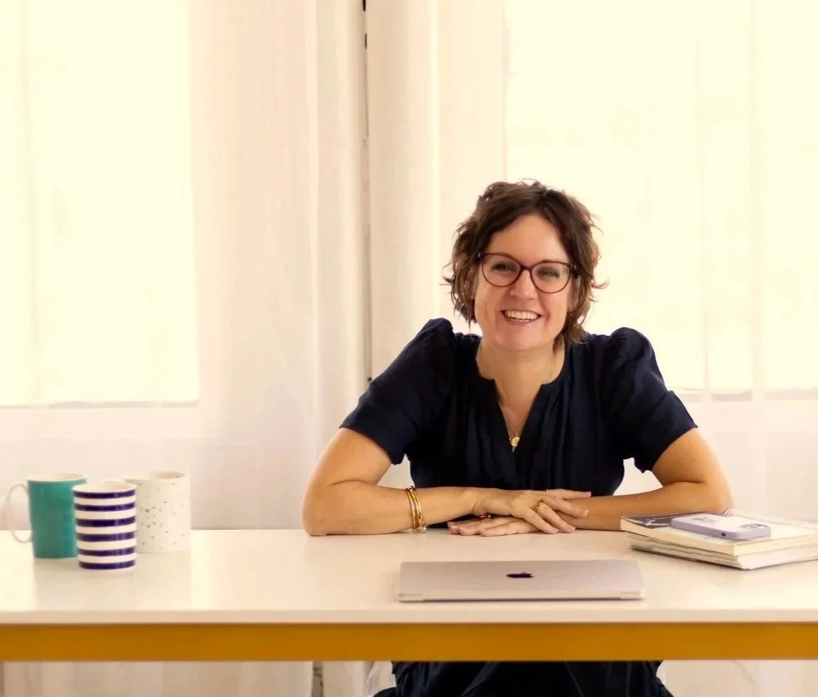 Woman with short brown hair, glasses, and wearing a black top, smiling and sitting at a white desk with a laptop, notebooks, and coffee mugs in a bright room with white curtains.