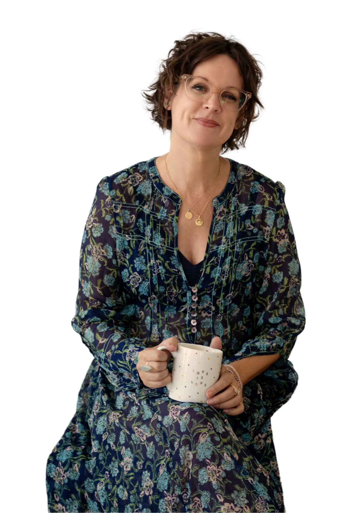 A woman with short curly hair and glasses, wearing a floral dress, holding a white mug that says 'Love is love', smiling softly.