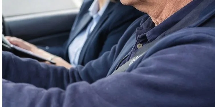 Close-up of a man in a navy jacket, sitting in a car, with a person in the background looking at a tablet.