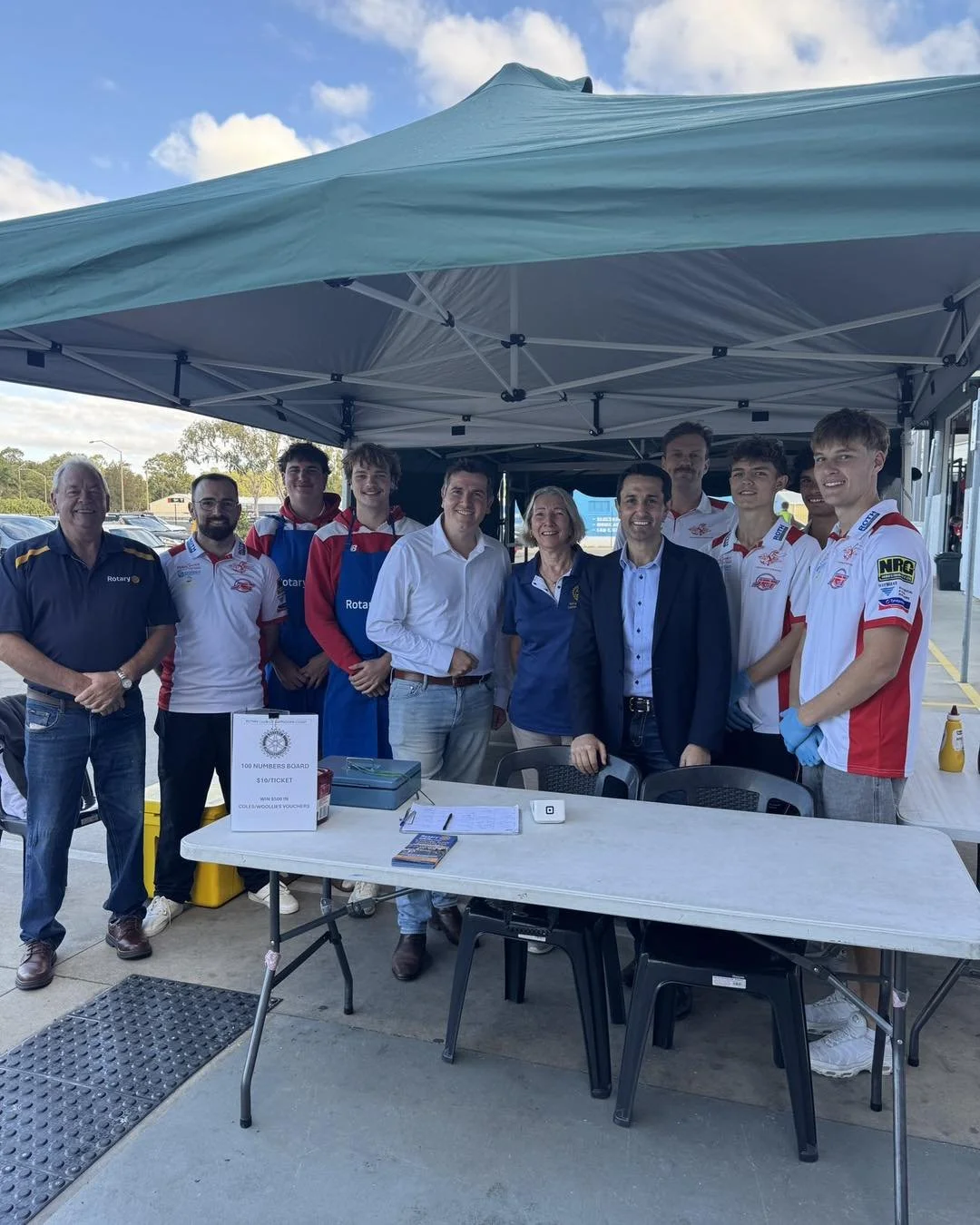 Image of a Bunnings Sausage Sizzle featuring Rotary Members, local and state politicians and the local under 18's AFL club. They are posing for the picture, looking at the photographer with their hands clasped in front of them.