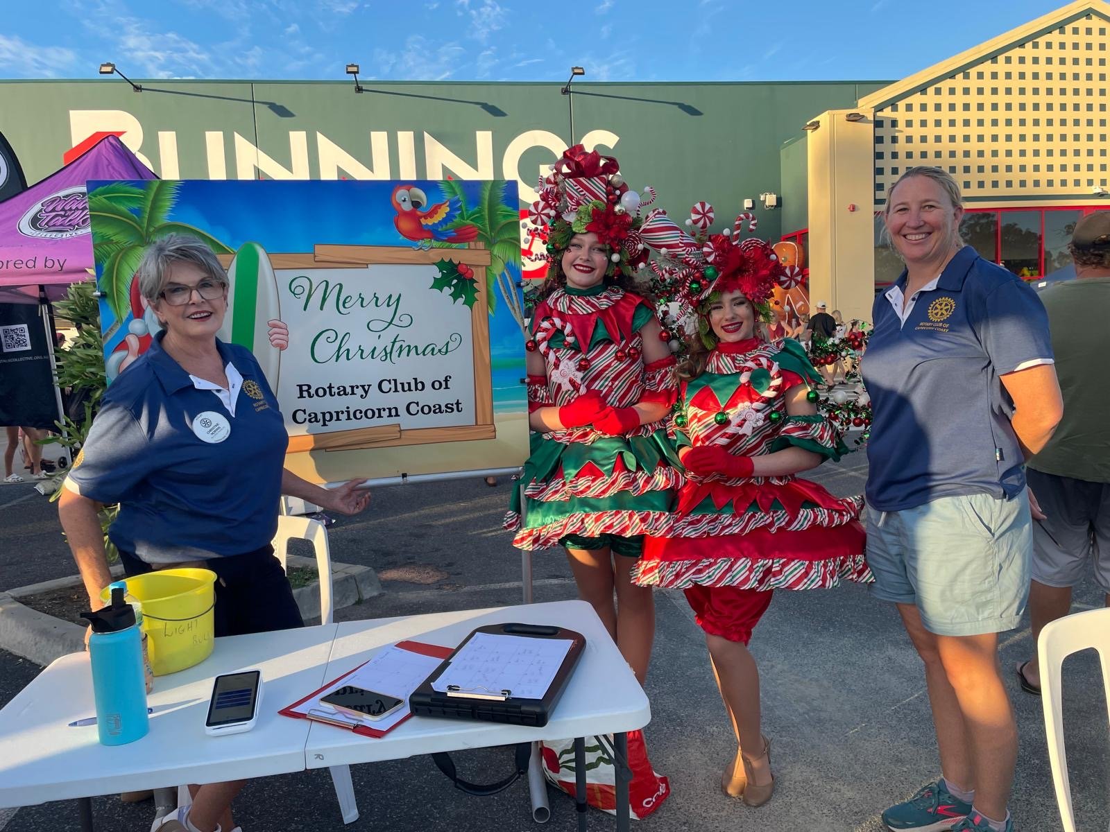 female Rotary members pose with two people dressed in Christmas costumes. The costumes are read and green with tinsel and baubles.