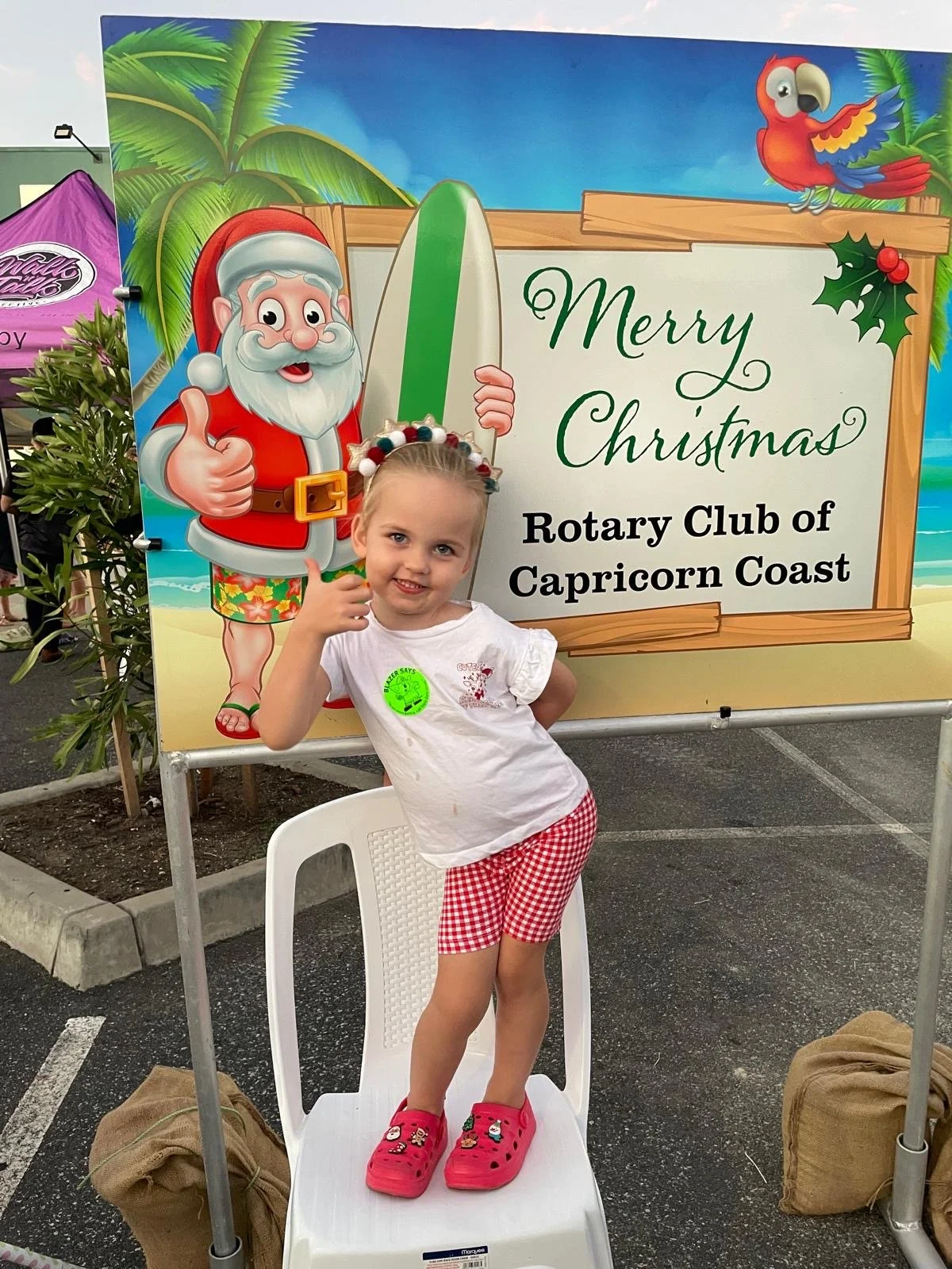 A young girl child poses in front of a Santa holding a surfboard. The sign reads - Merry Christmas Rotary Club of Capricorn Coast