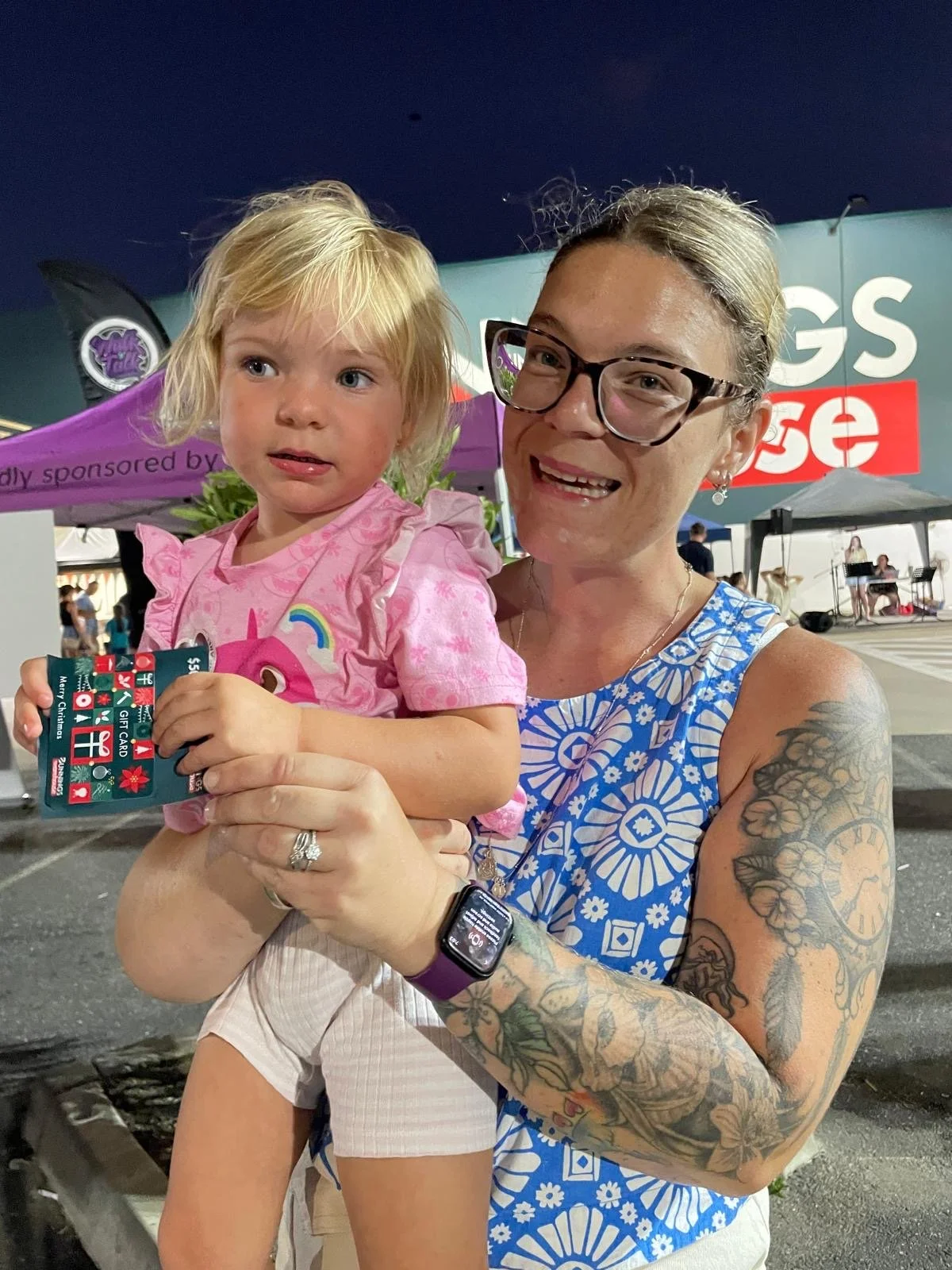 A young mom holds up her toddler daughter. Both are smiling. The toddlers holds up a Bunnings gift certificate