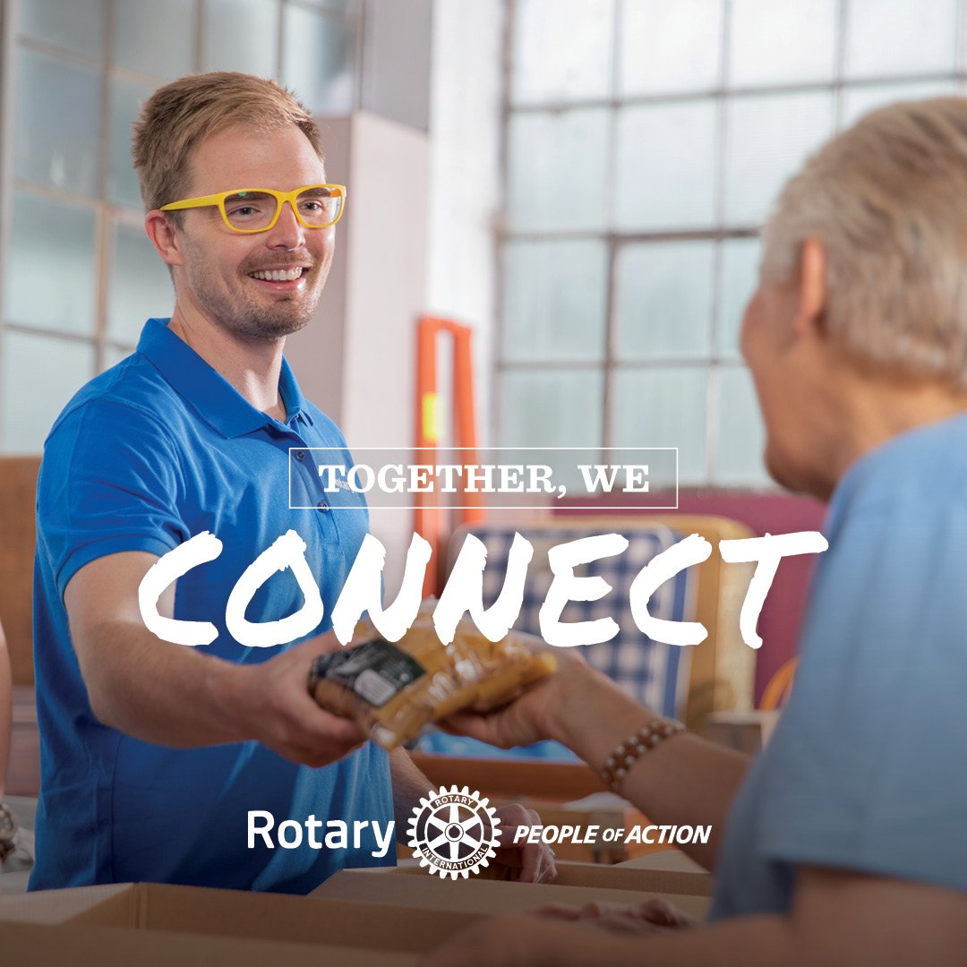 A young man wearing a Rotary shirt and glasses is handing a loaf of bread to a senior.