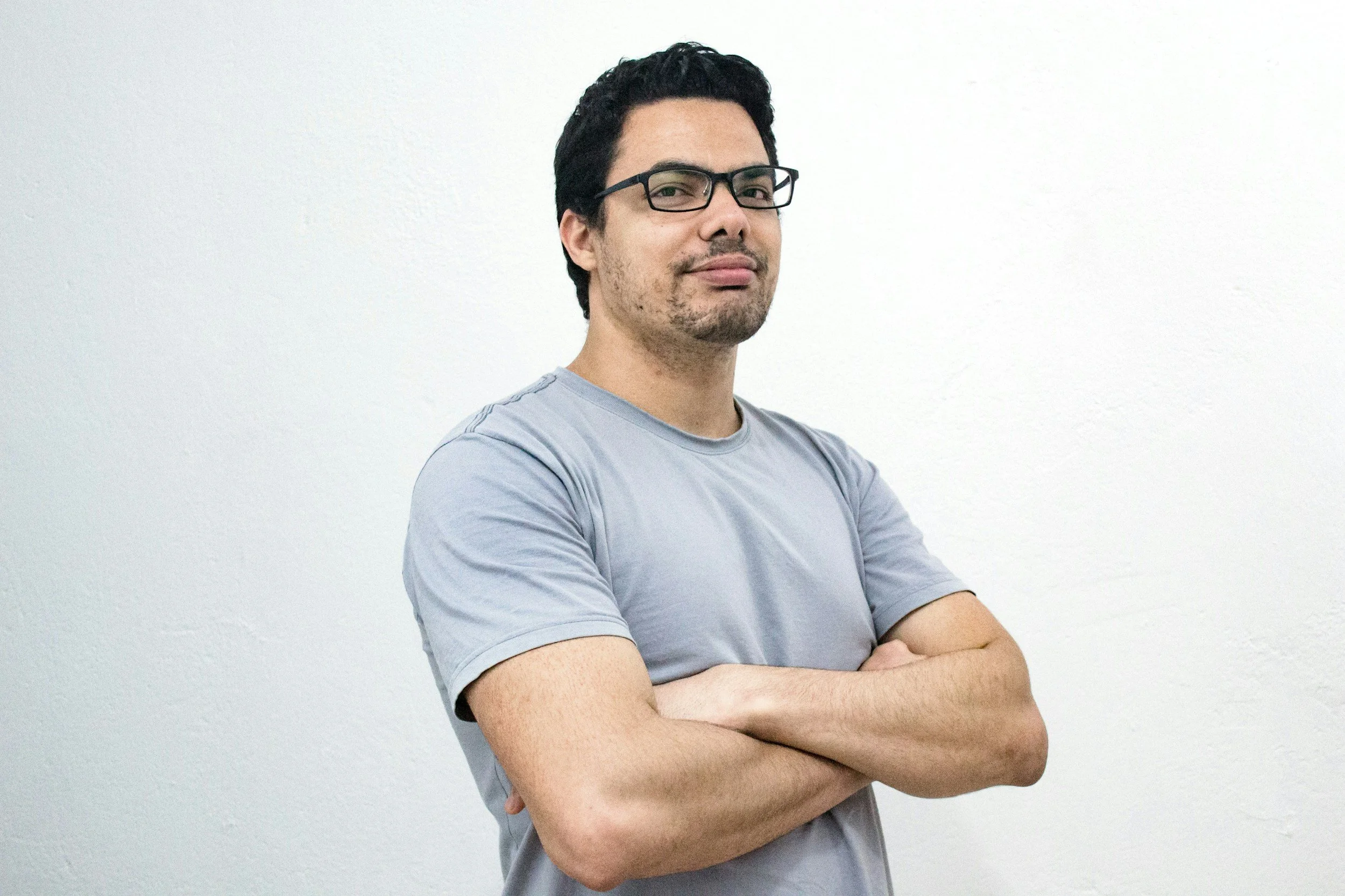 A man with glasses, dark hair, and a light beard wearing a light gray t-shirt, crossing his arms, standing against a plain white wall.