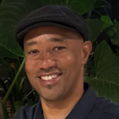 A smiling man wearing a gray cap and dark shirt, standing in front of green large-leafed plants.