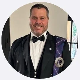 Man in formal attire with a black tuxedo, white dress shirt, and black bow tie, smiling indoors, with a window in the background.