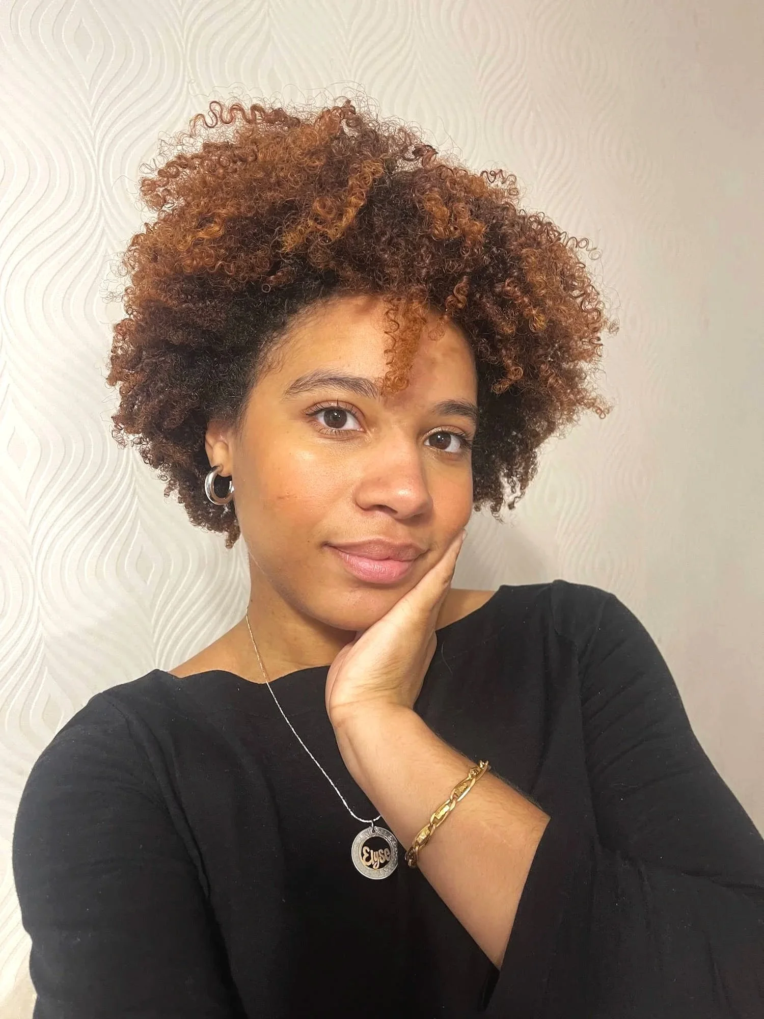 Close-up of a woman with short, curly, reddish-brown hair, wearing earrings, a black top, and gold jewelry, posing indoors against a textured, off-white wall.