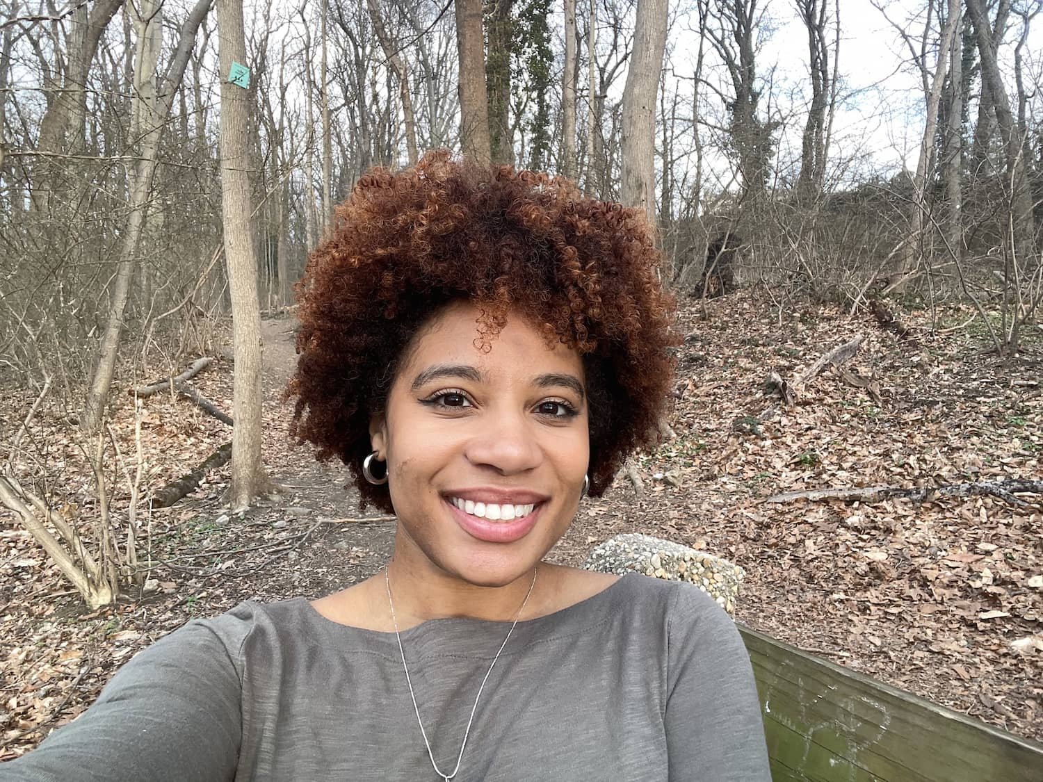 Smiling woman with curly hair taking a selfie outdoors in a wooded area with fallen leaves on the ground.