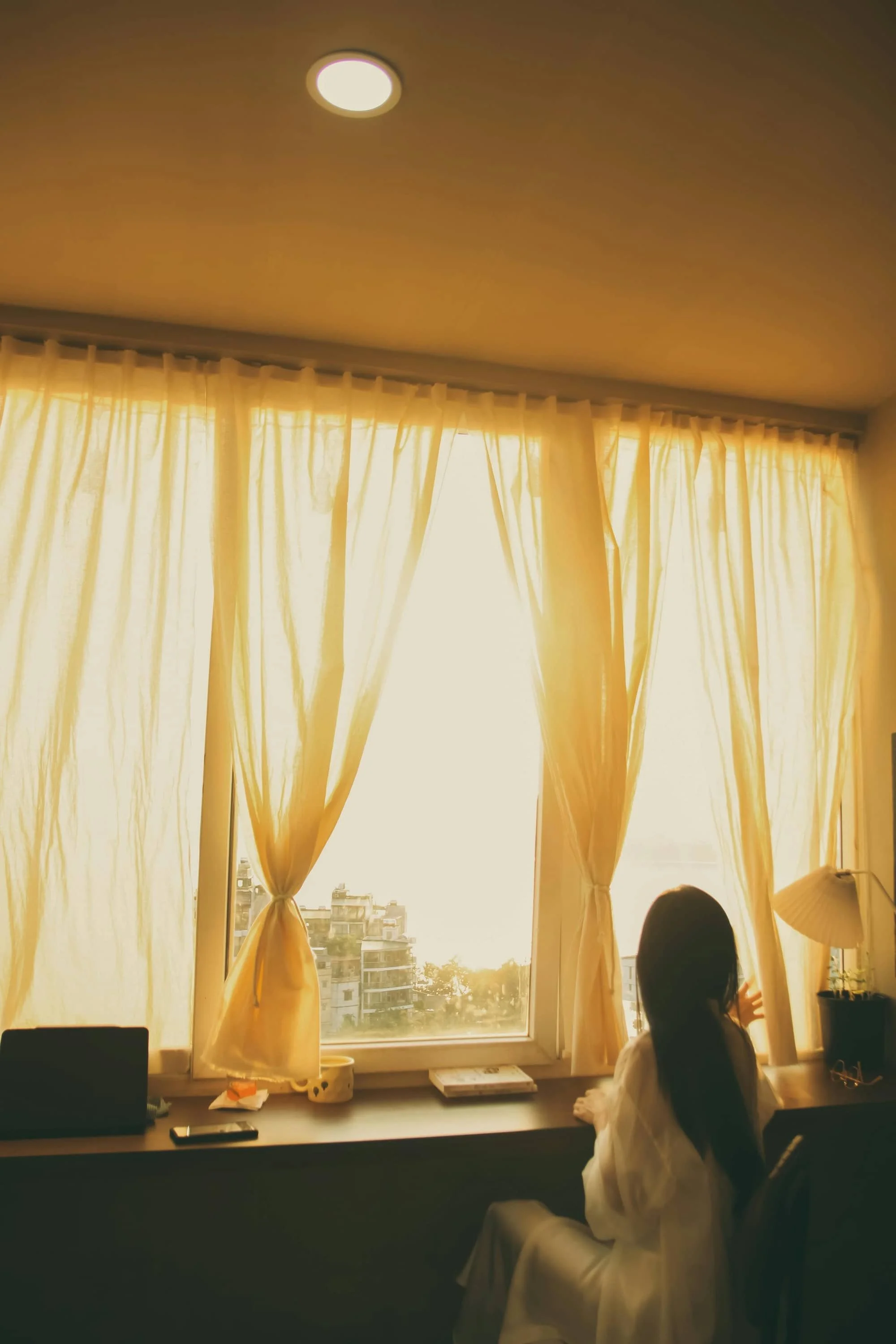 A girl in a white dress sitting on a desk by a window with yellow curtains, looking outside at buildings during sunset or sunrise.